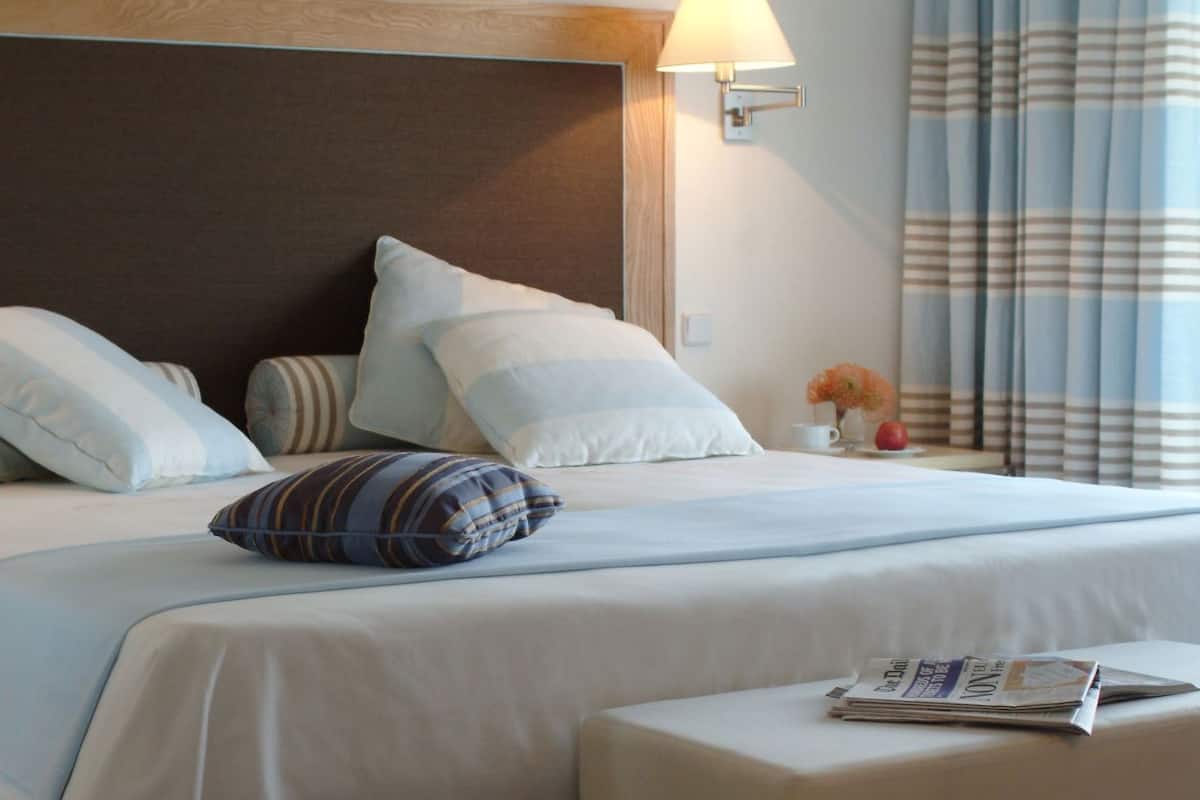 Habitación de hotel con cama, almohadas, mesita de noche, lámpara, ventana con cortinas y alfombra.