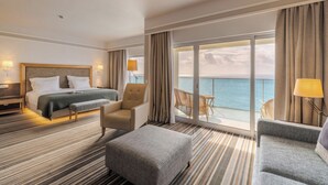Une chambre d’hôtel comprenant un lit, deux fauteuils, un canapé, un banc, un balcon avec vue sur la mer et une grande fenêtre.