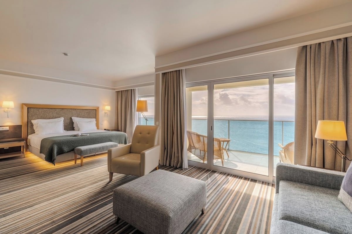 Habitación de hotel con cama, dos sillones, un sofá, un banco, balcón con vistas al mar y un ventanal grande.