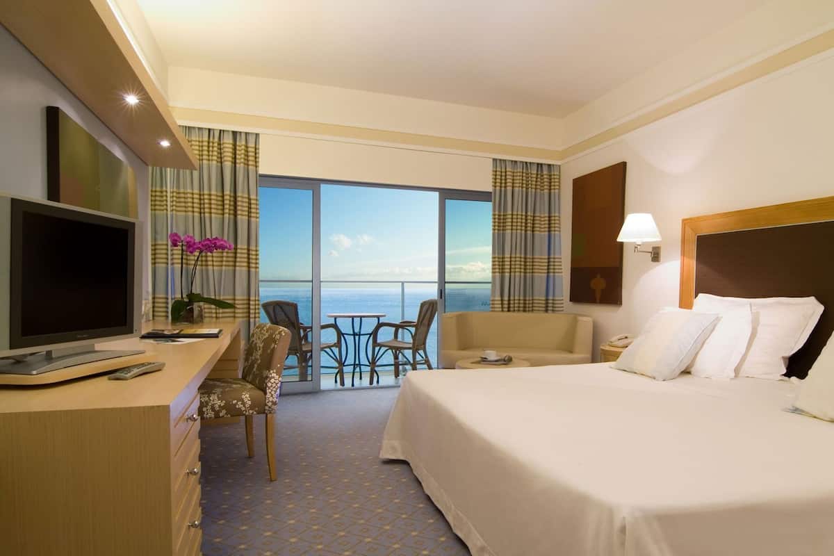 Habitación de hotel con cama, escritorio con televisor, silla, una mesita y vista al océano.