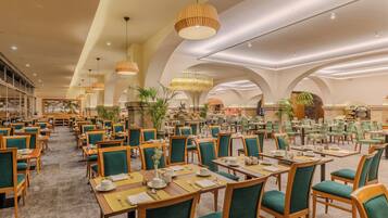 Un vaste espace dédié à la salle à manger, avec de nombreuses tables et chaises, un lustre décoratif central et une grande fenêtre offrant une vue panoramique.