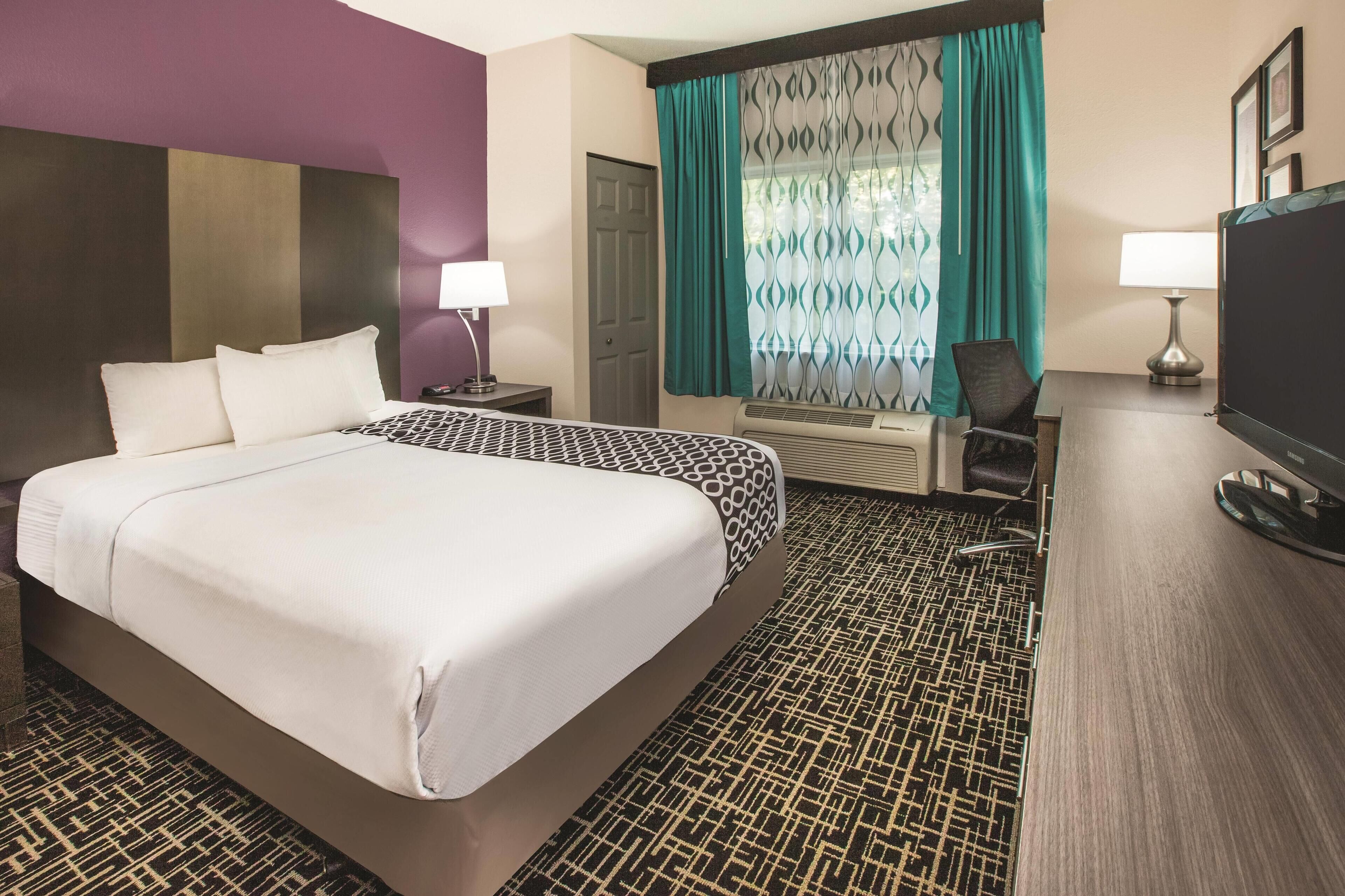A hotel room with a large bed, a desk with a computer, a chair, a TV, and a window with curtains.