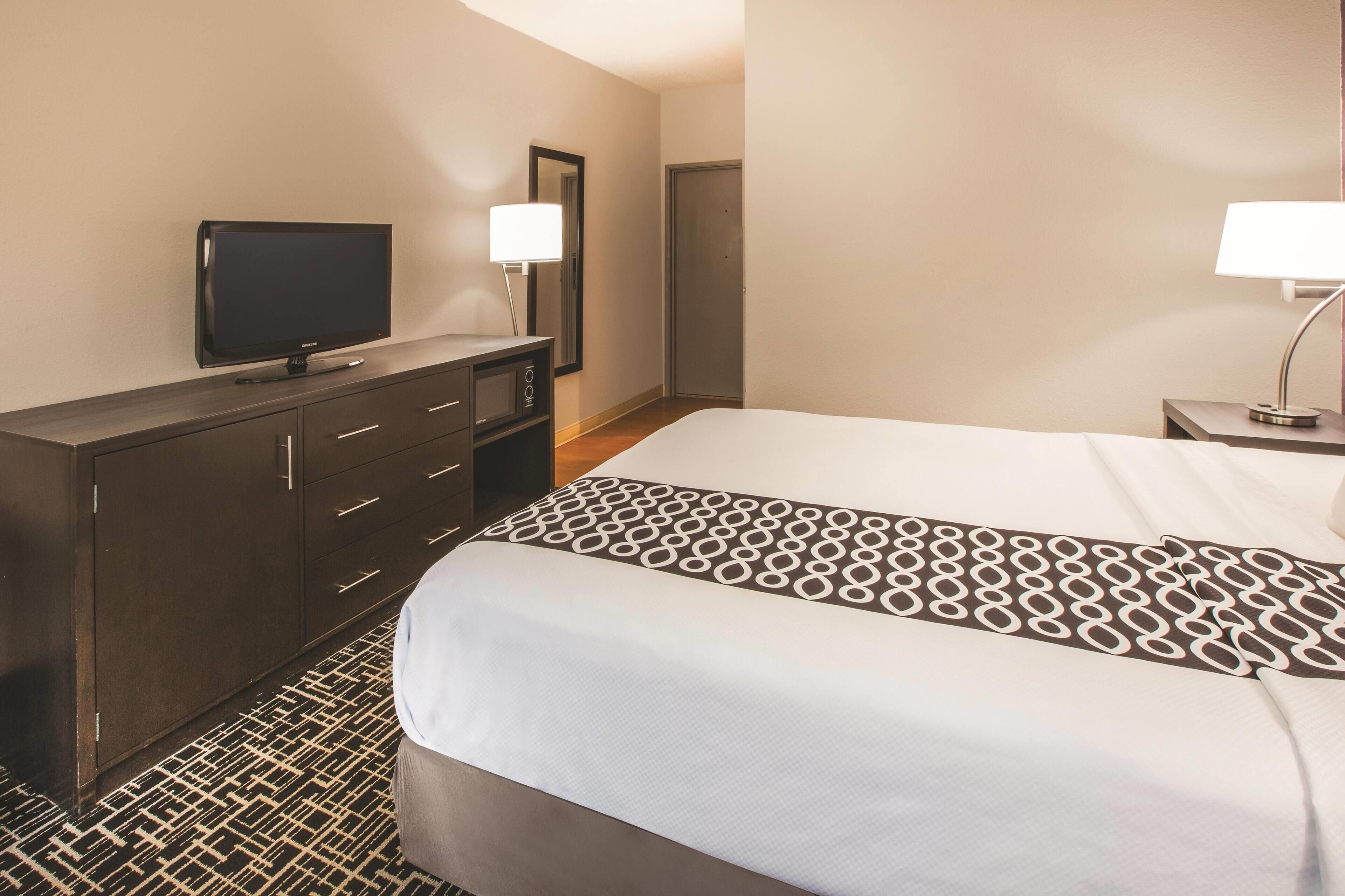 A hotel room with a bed, a television on a dresser, a mirror, and a nightstand with a lamp.