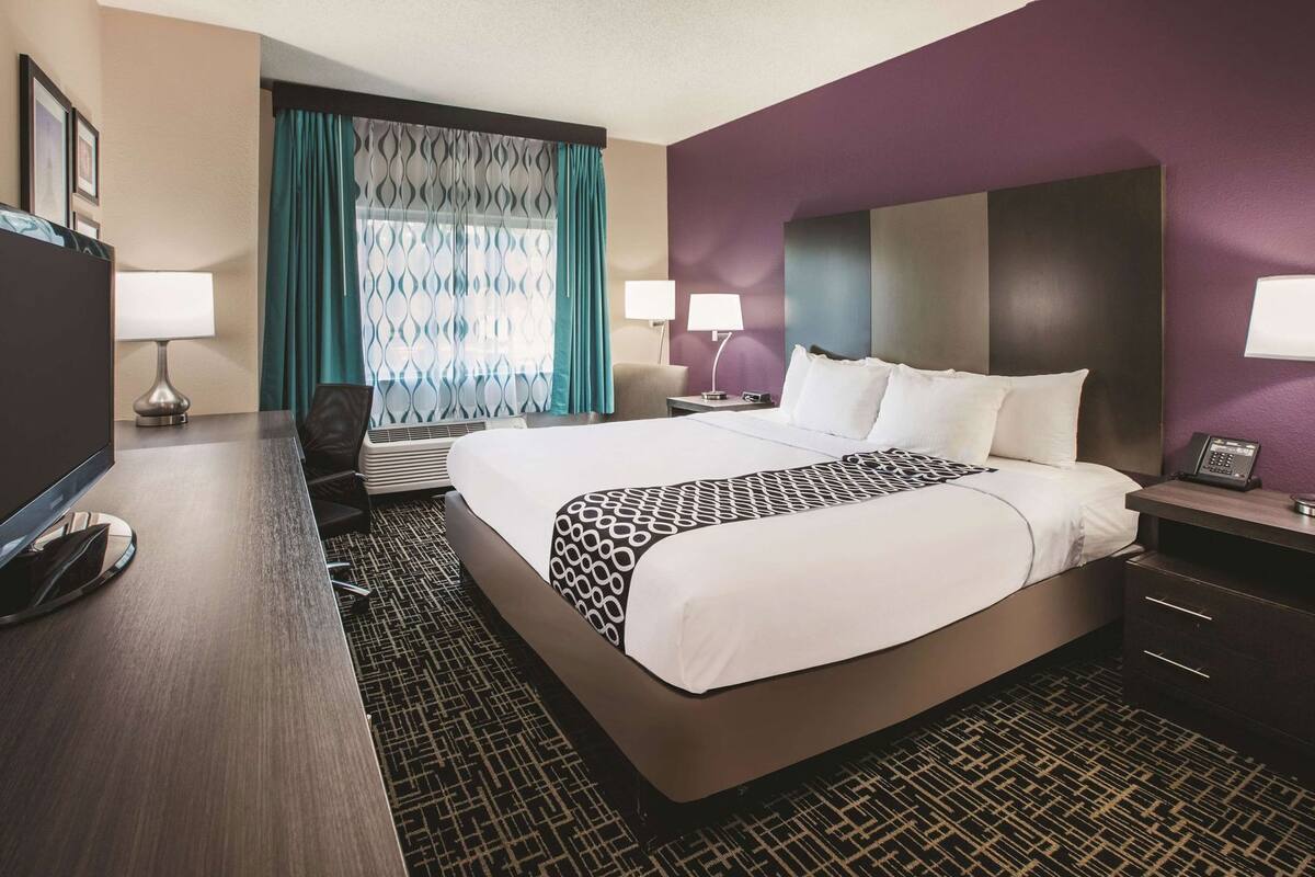 A hotel room with a large bed, two bedside tables, a desk with a chair, a TV, and a window with curtains.