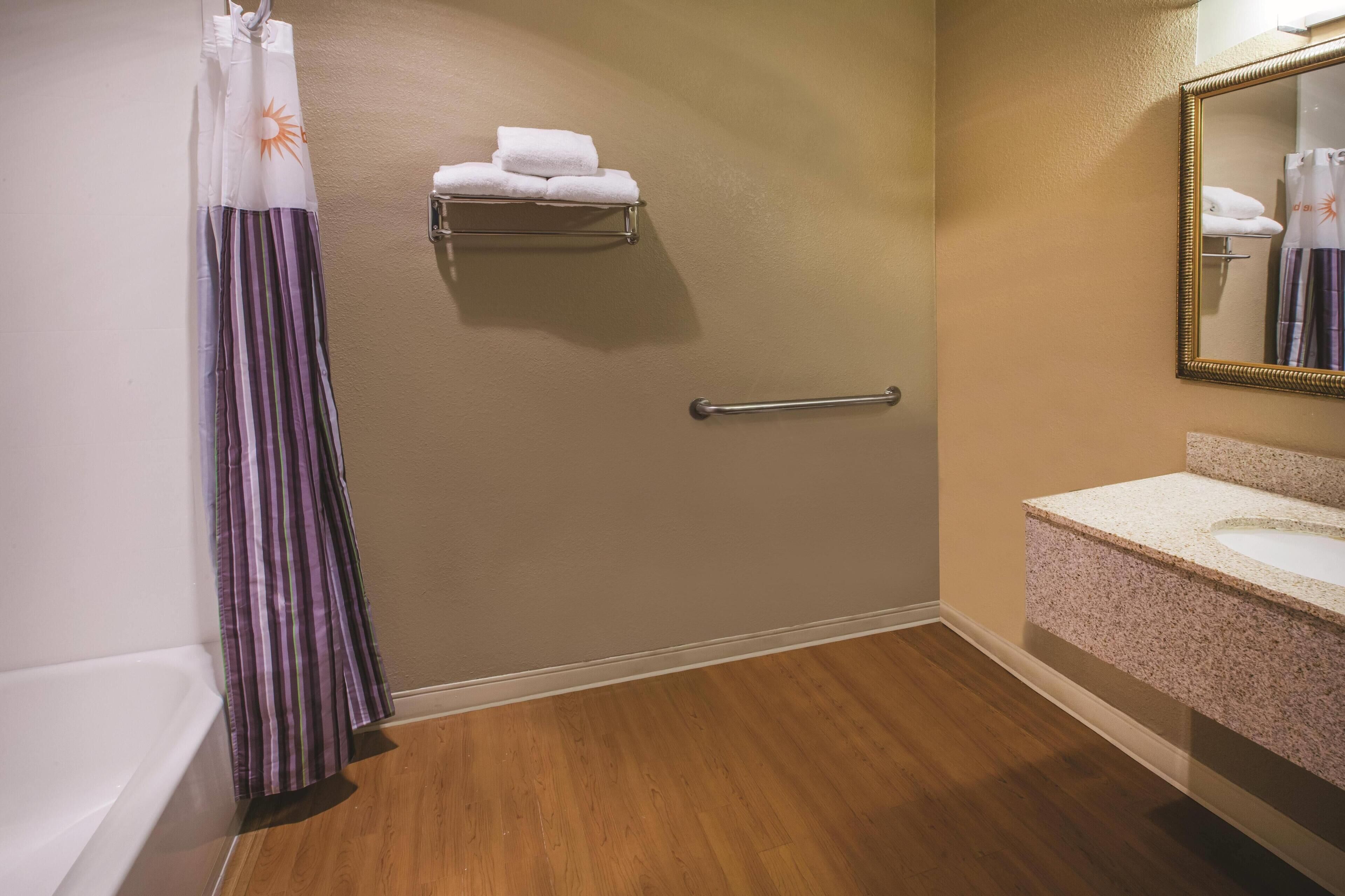 A bathroom with a shower curtain, a towel rack with towels, a handrail, a sink with a mirror, and wooden flooring.
