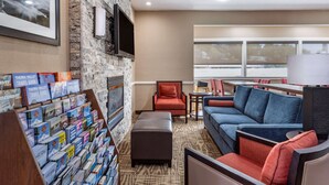 A hotel lobby with a stone wall, a mounted TV, a seating area with a blue sofa, a red armchair, and a bookshelf with travel guides. - Comfort Inn Kelso - Longview (Kelso)