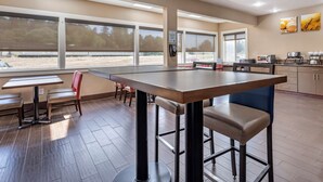A modern dining area with a long table and chairs, large windows, and a kitchen area with appliances. - Comfort Inn Kelso - Longview (Kelso)