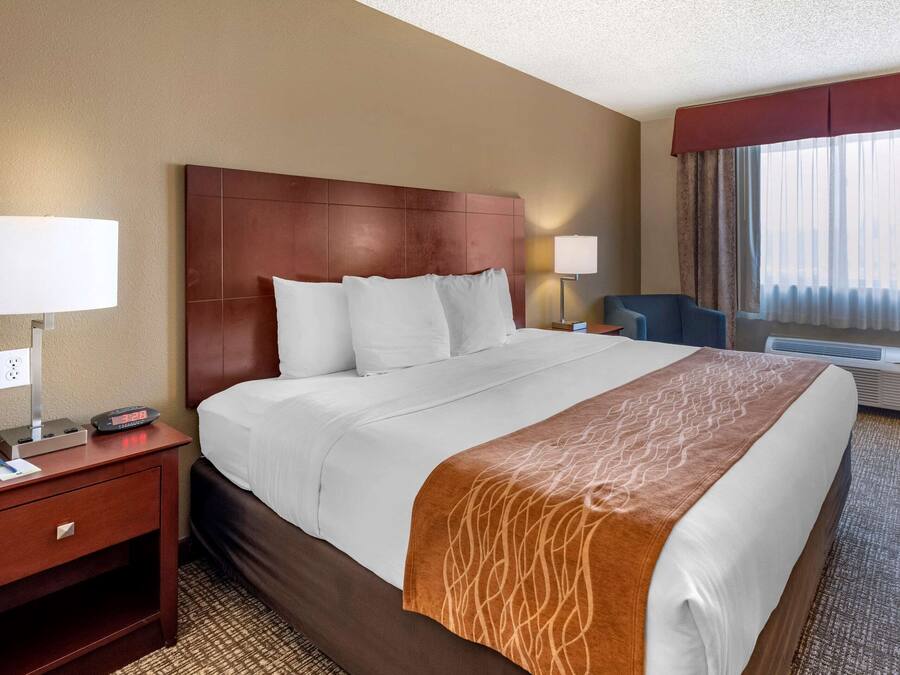 A hotel room with a large bed, a wooden headboard, two bedside lamps, a desk with a telephone, and a window with curtains.
