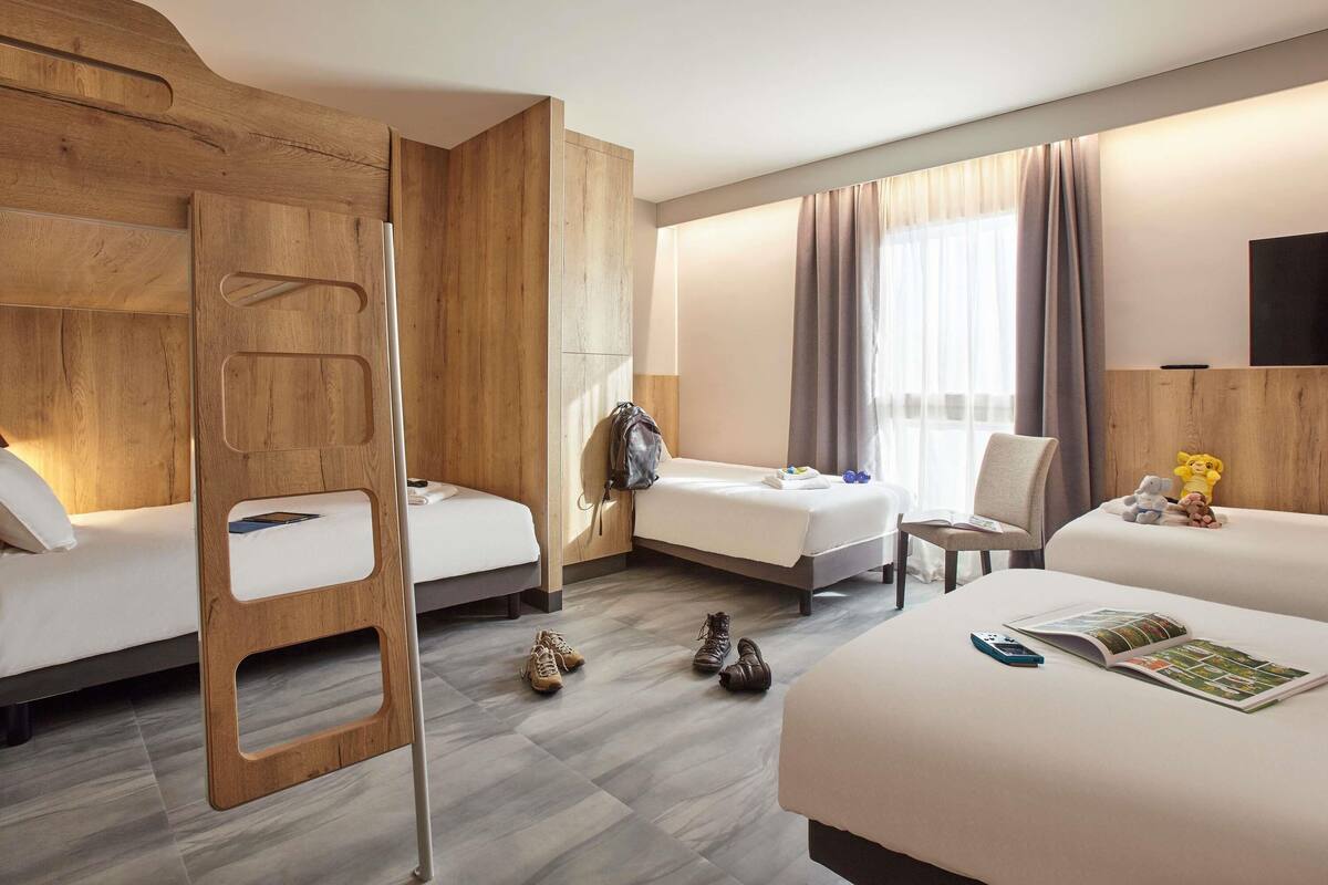 A hotel room with a bunk bed, a single bed, a chair, a TV, and a window with curtains.