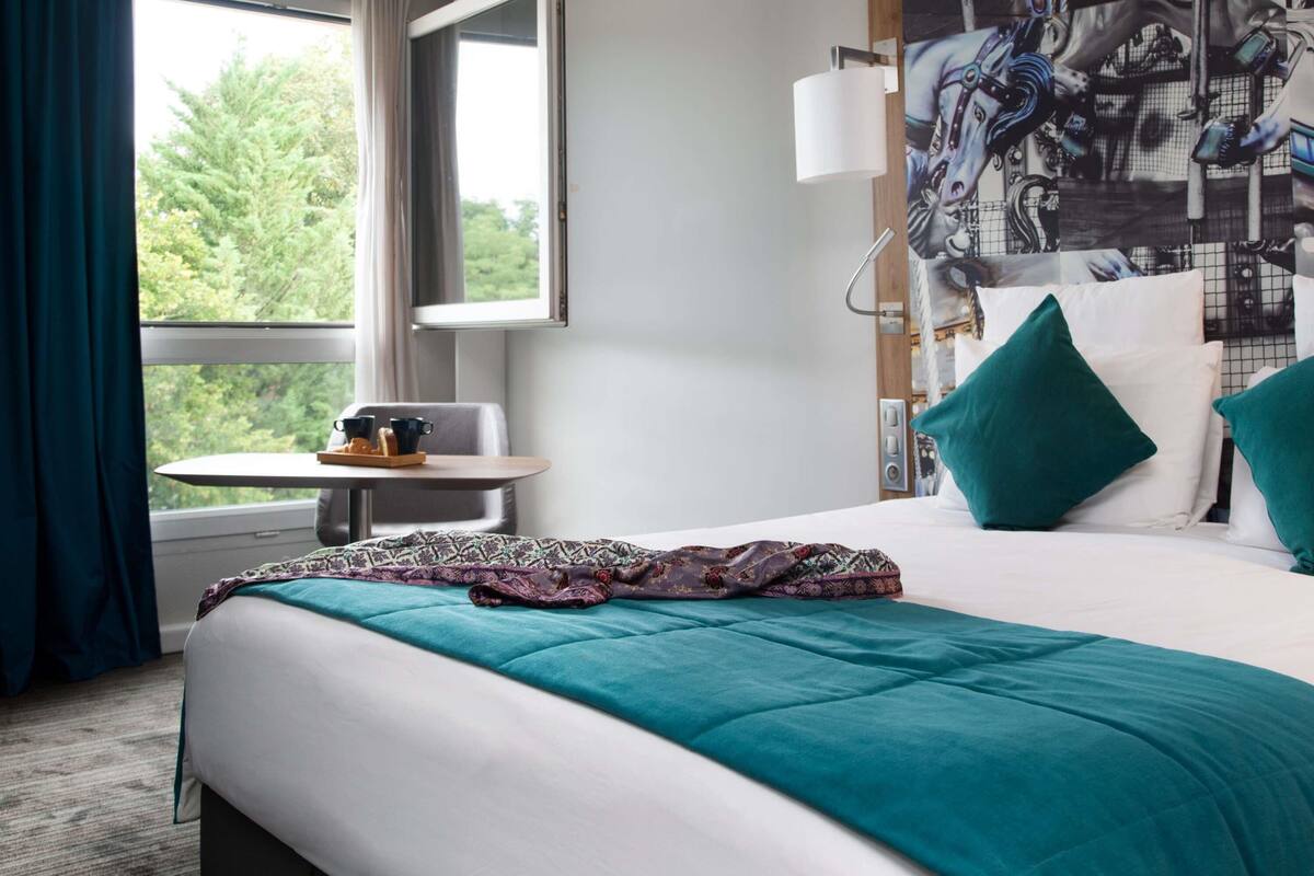 A hotel room with a bed, a small table with a tray of items, a window with curtains, and a wall with a large abstract painting.