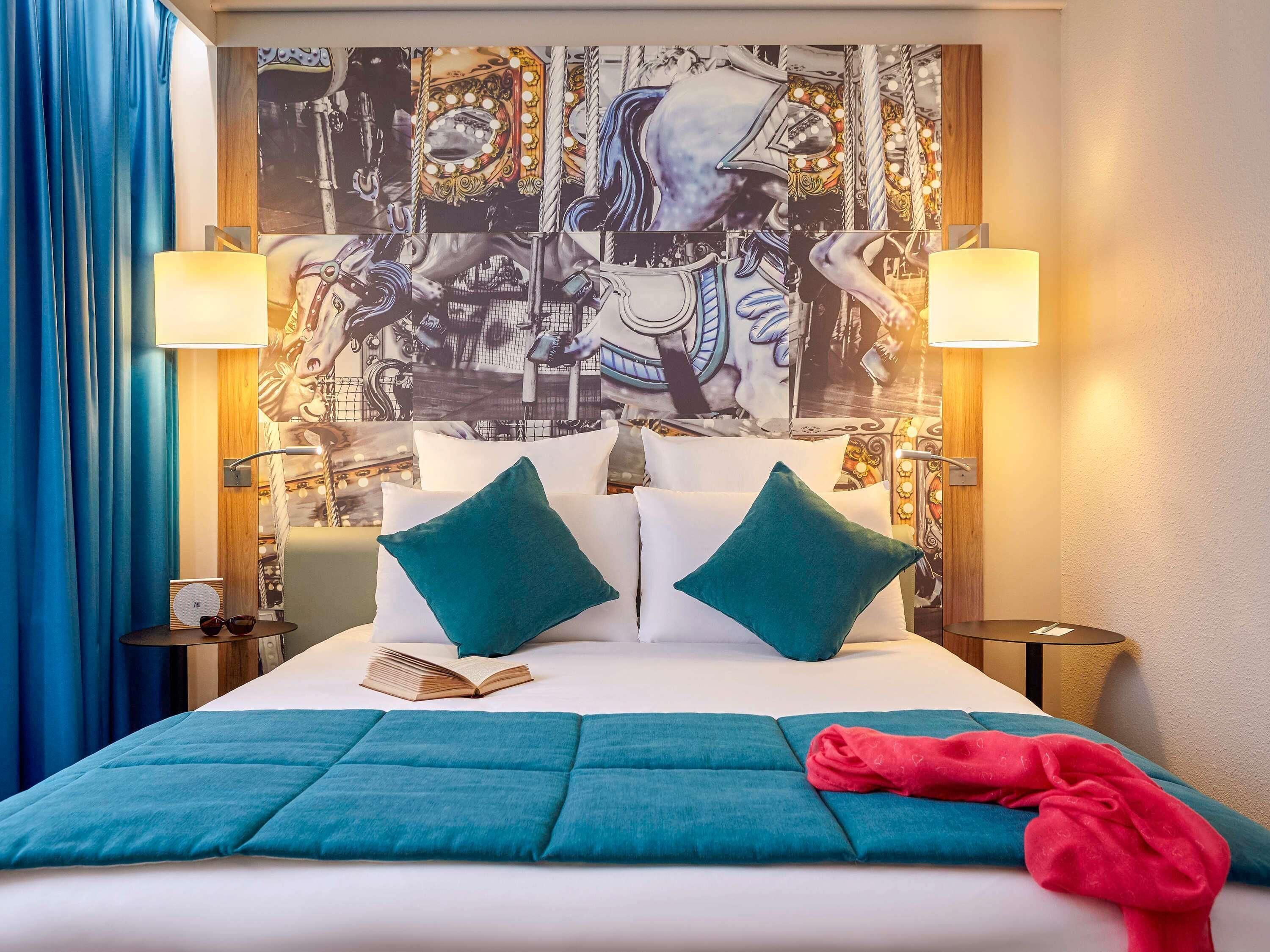 A hotel room with a large bed, two teal pillows, a red blanket, and a wall mural.