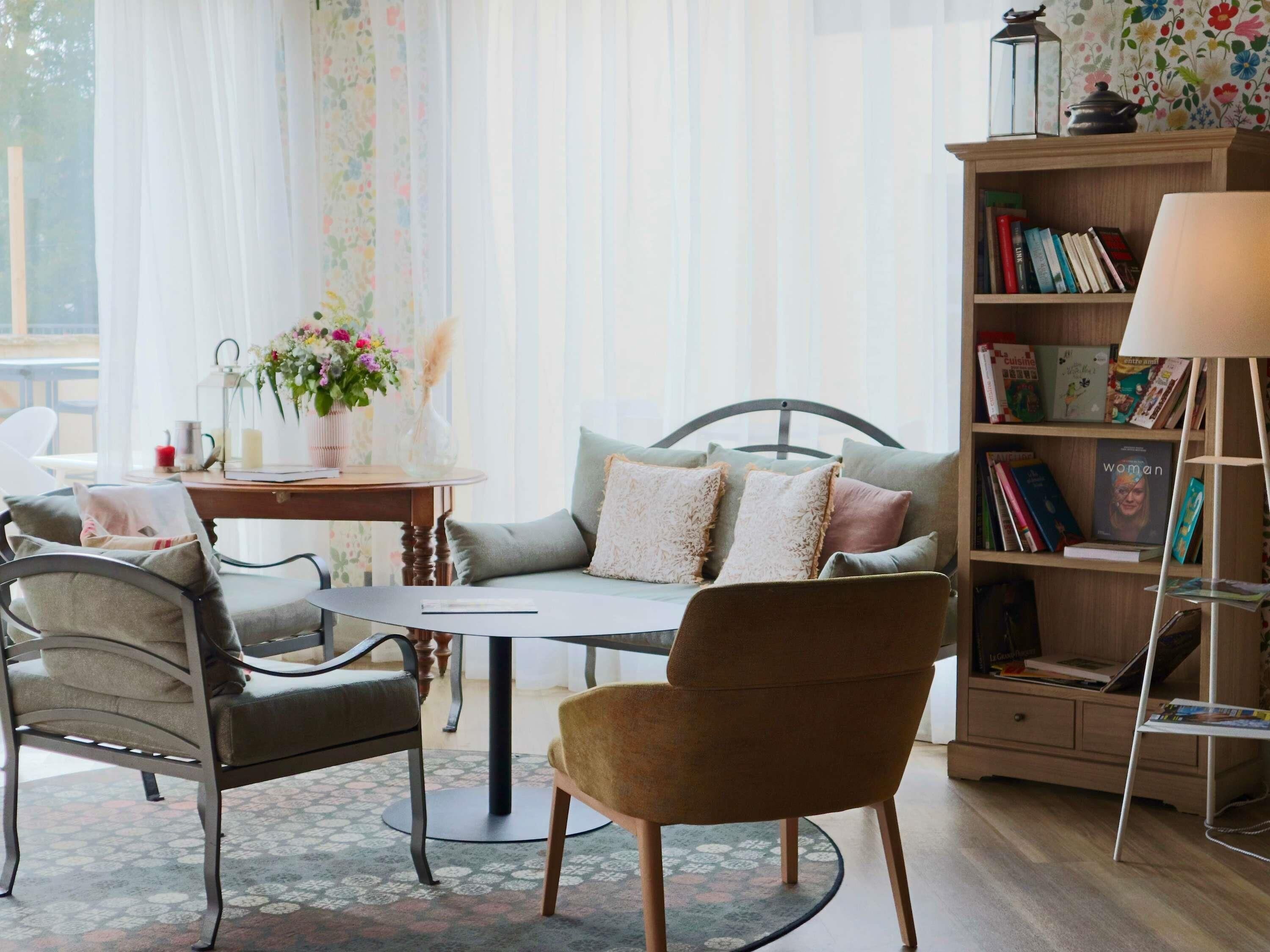 Una sala de estar acogedora con un sofá, sillas, una mesa de centro, una estantería y una ventana con cortinas traslúcidas.