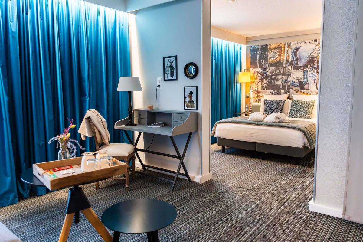 A hotel room with a desk, chair, bed, and a view of the cityscape.