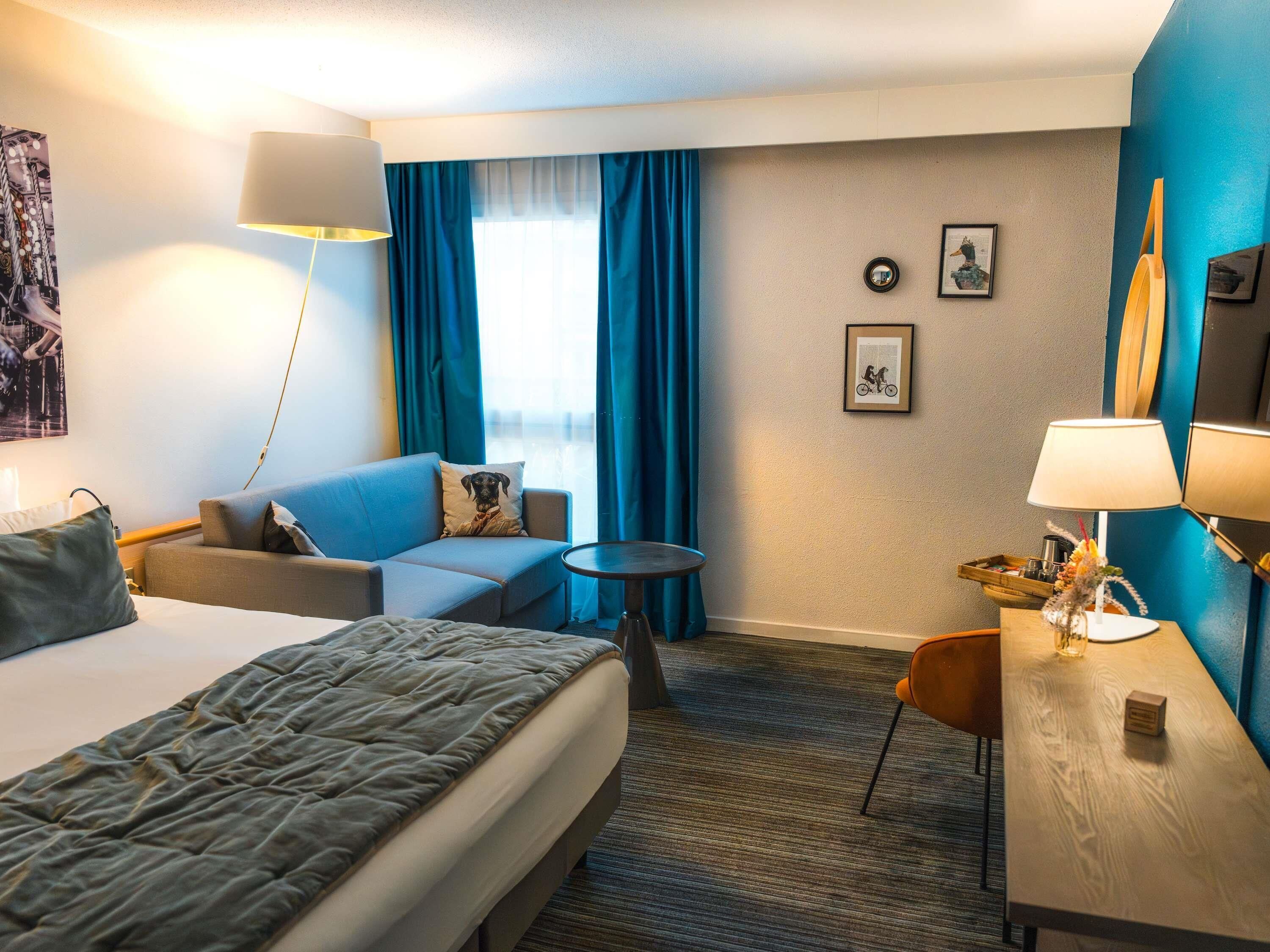 A hotel room with a bed, a sofa, a small table, a desk, a lamp, and a view of the outdoors.