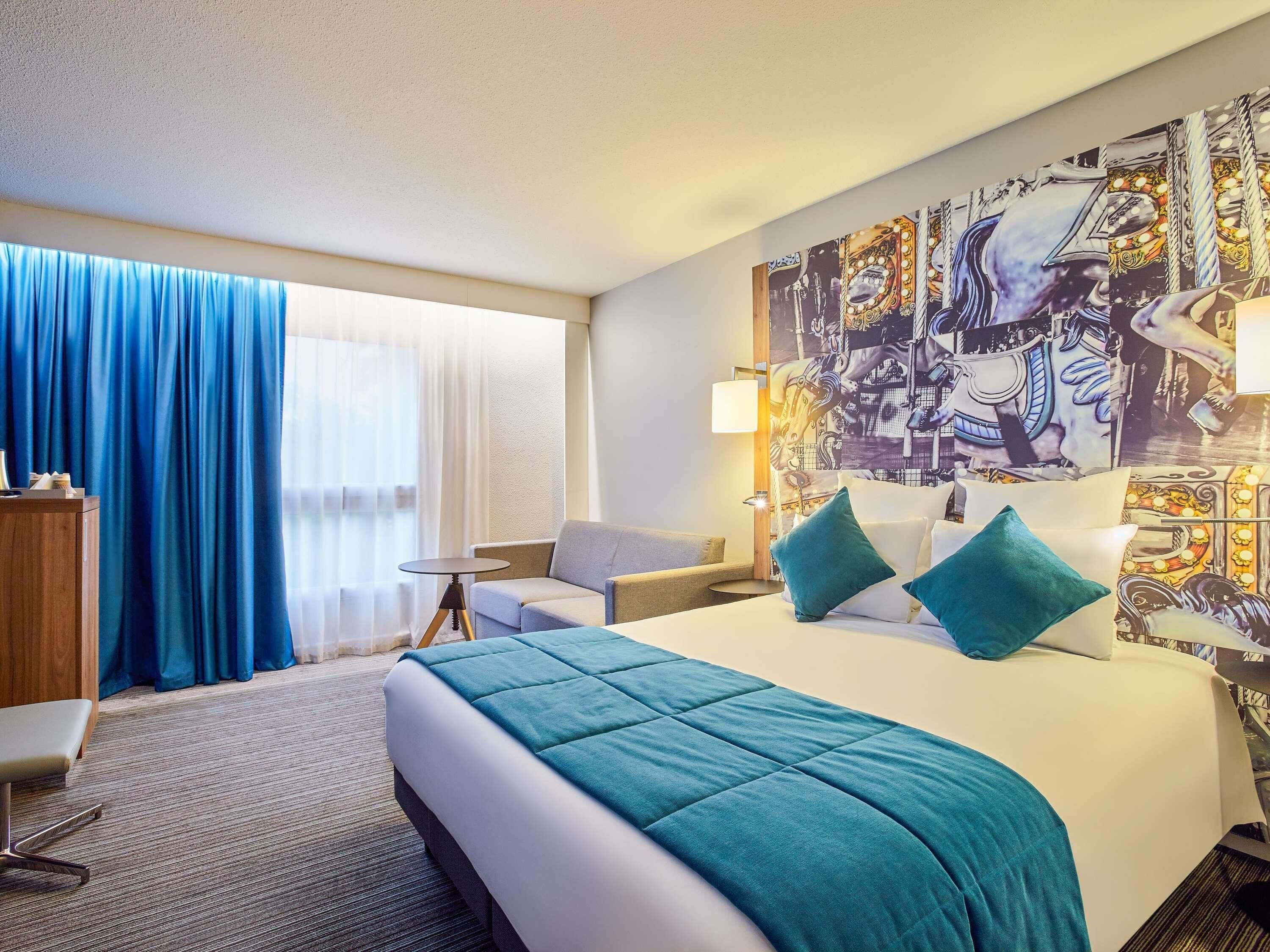 Habitación de hotel con cama, escritorio, silla, ventana grande con cortinas y pared con un mural.