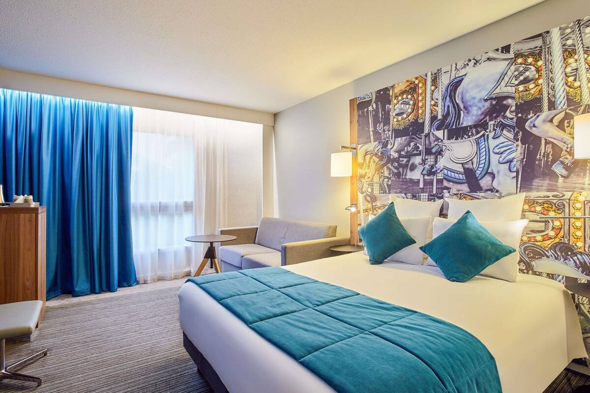 A hotel room with a bed, a desk, a chair, a large window with curtains, and a wall with a mural.