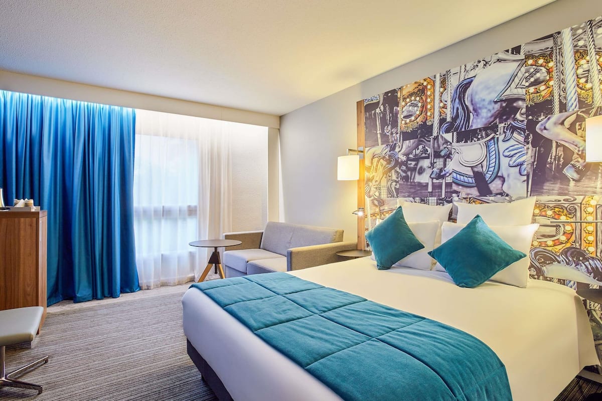 Habitación de hotel con cama, escritorio, silla, ventana grande con cortinas y pared con un mural.