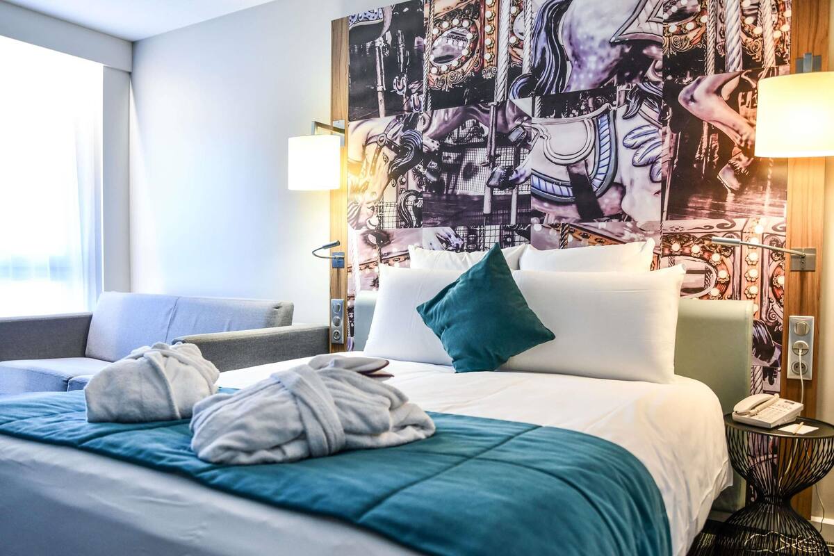 A hotel room with a bed, two bedside lamps, a telephone, and a wall with a large poster.