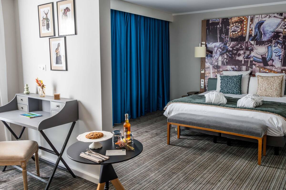A hotel room with a bed, a desk with a chair, a small table with a cookie and drinks, and a blue curtain.