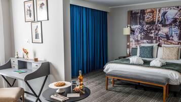 A hotel room with a bed, a desk with a chair, a small table with a cookie and drinks, and a blue curtain.