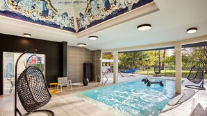 A modern indoor pool area with a clear view of the outdoors, a seating area with chairs, and a decorative ceiling mural. - Demeures de campagne Château de Fontainebleau – Mercure (Fontainebleau)