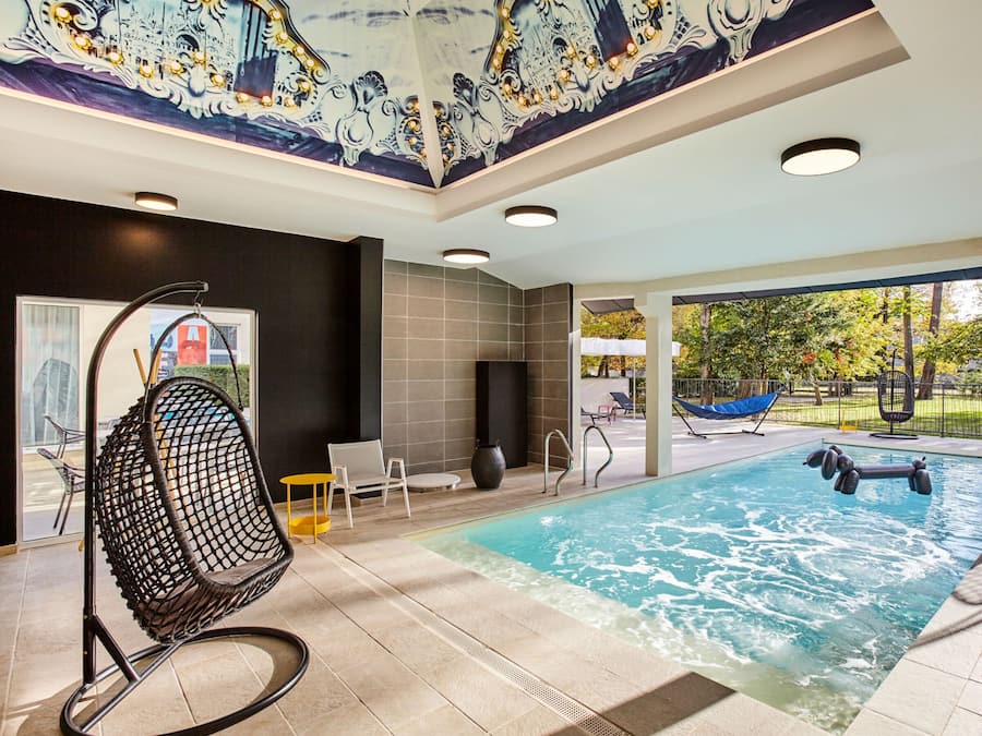 A modern indoor pool area with a clear view of the outdoors, a seating area with chairs, and a decorative ceiling mural.