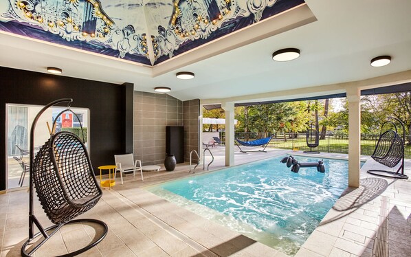 Un área de piscina interior moderna con vistas despejadas al exterior, una zona de estar con sillones y un mural decorativo en el techo.