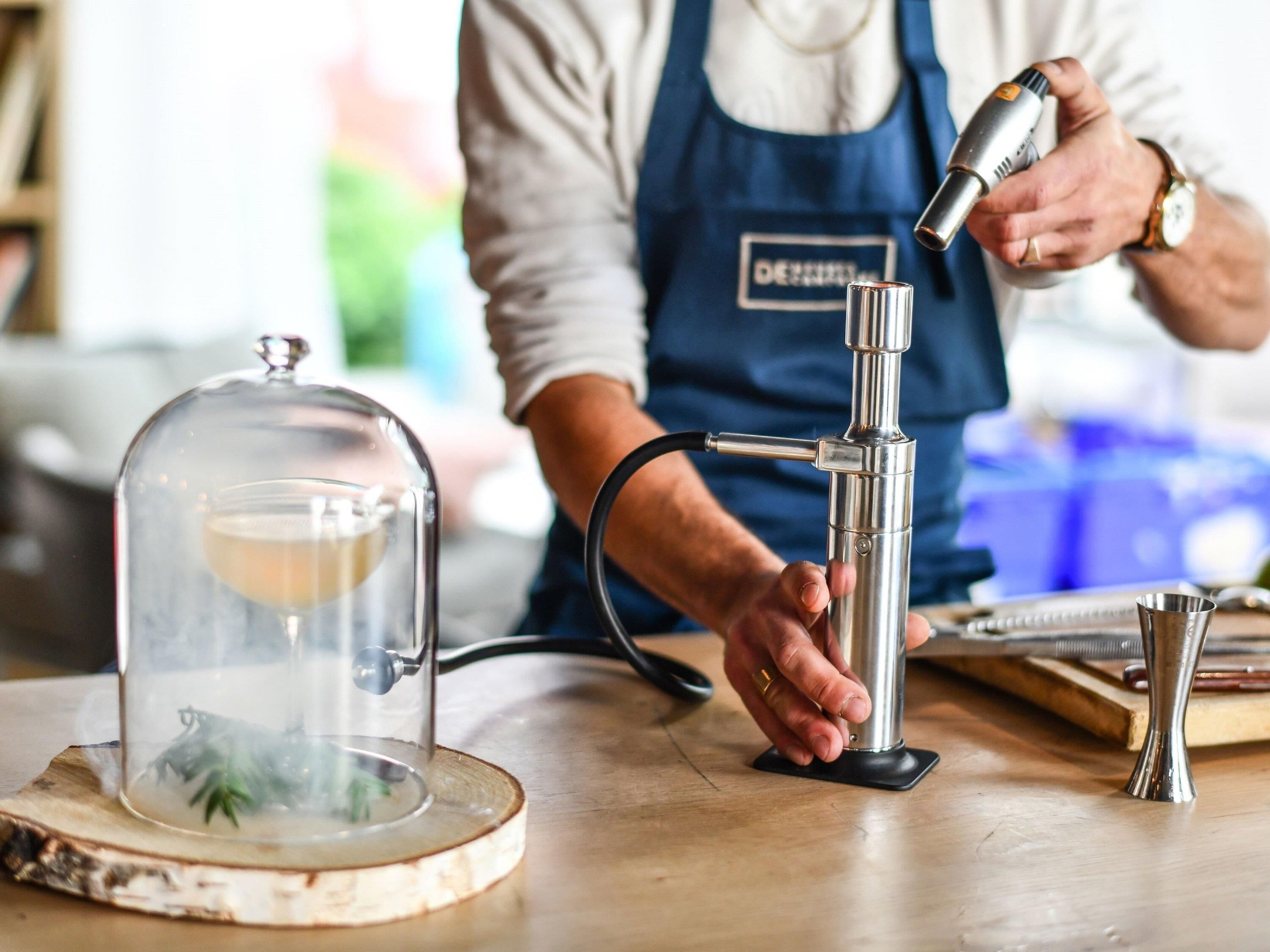 Una persona con delantal azul operando una máquina de café, con una jarra de vidrio y una jarra de metal sobre el mostrador.