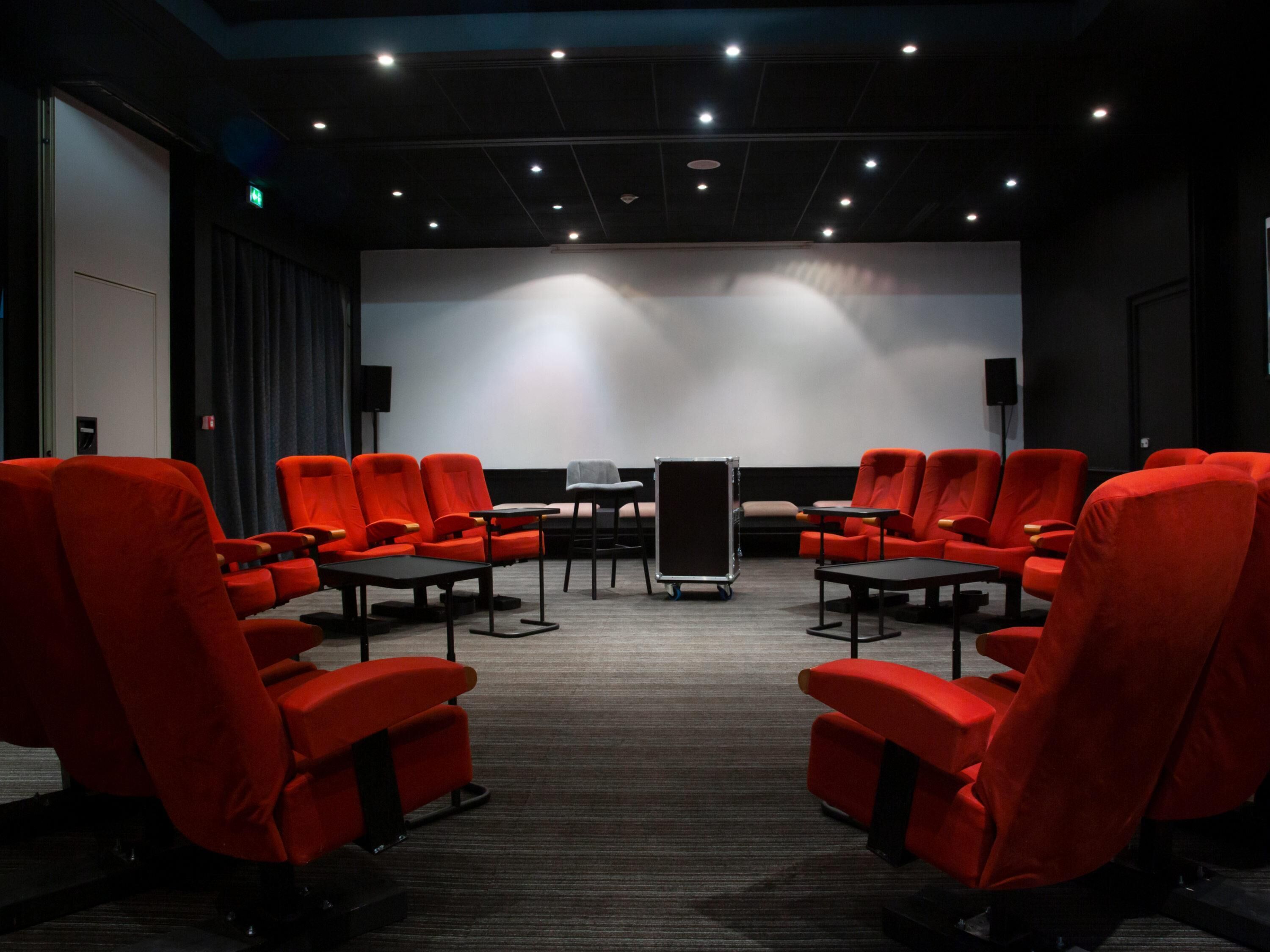 A conference room with rows of red chairs, a projector screen, and a speaker on the wall.