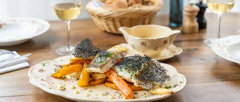 A dining table with a plate of fish, fries, and a decorative net on top. There are glasses of white wine and a basket of bread in the background.