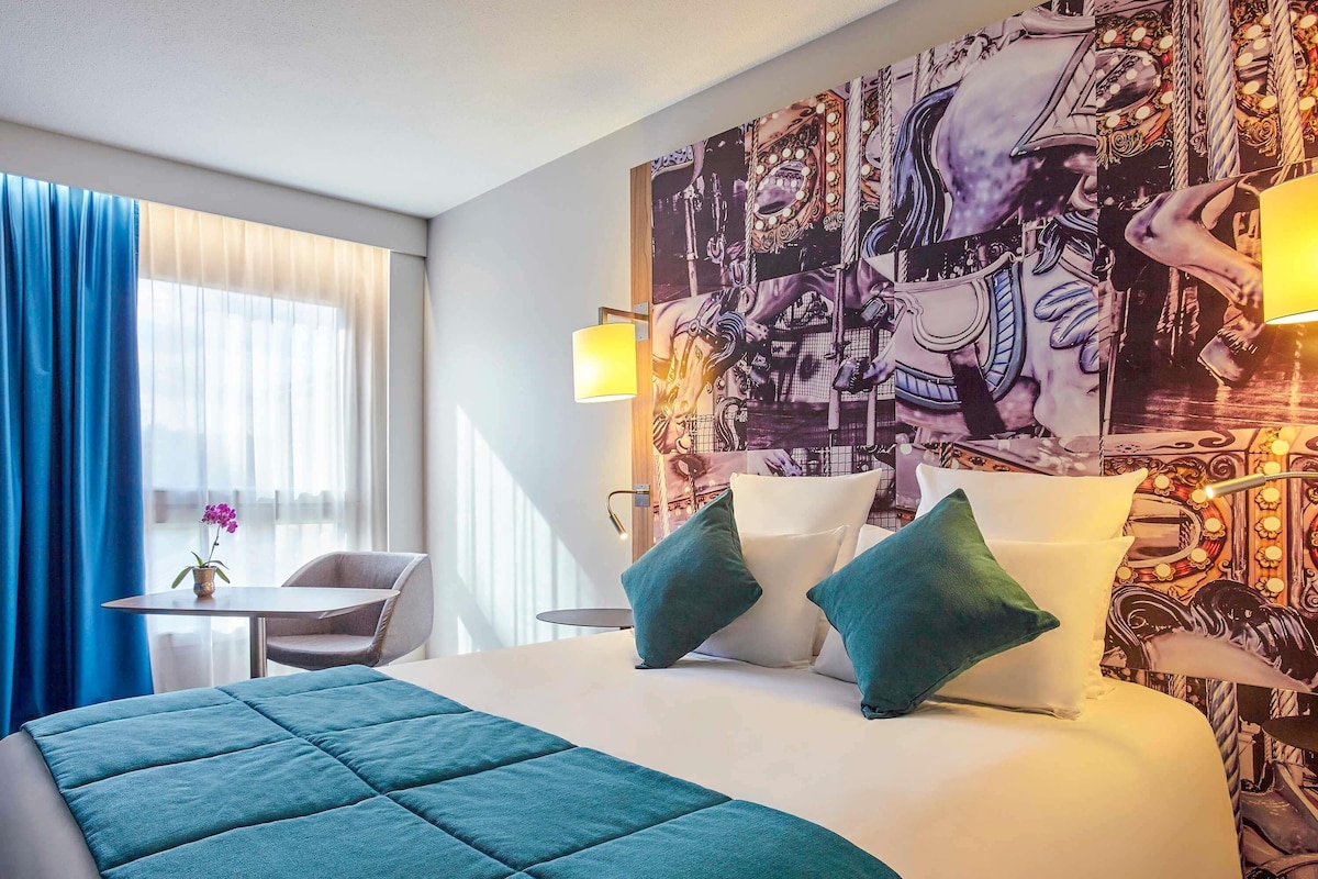 Een hotelkamer met een groot bed, twee kussens, een tafeltje met een vaas, en een stoel.