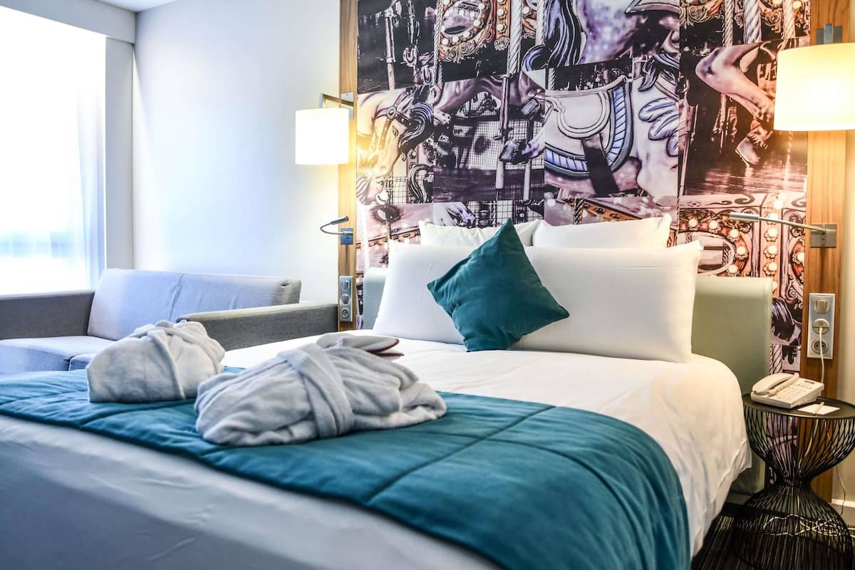 A hotel room with a bed, two bedside lamps, a telephone, and a wall with a large poster.