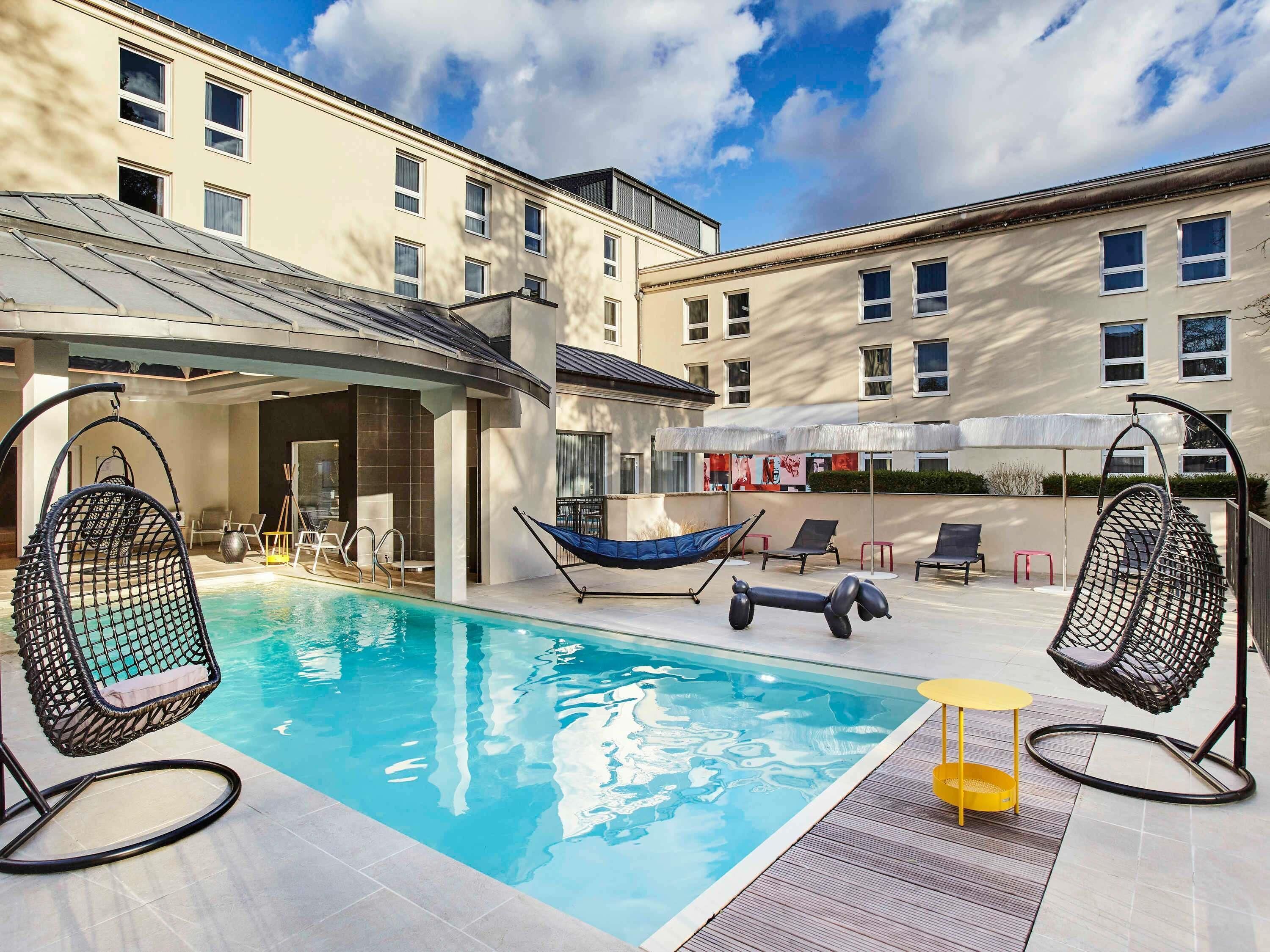 A swimming pool with lounge chairs and a hammock, surrounded by a building with multiple windows and a covered seating area.