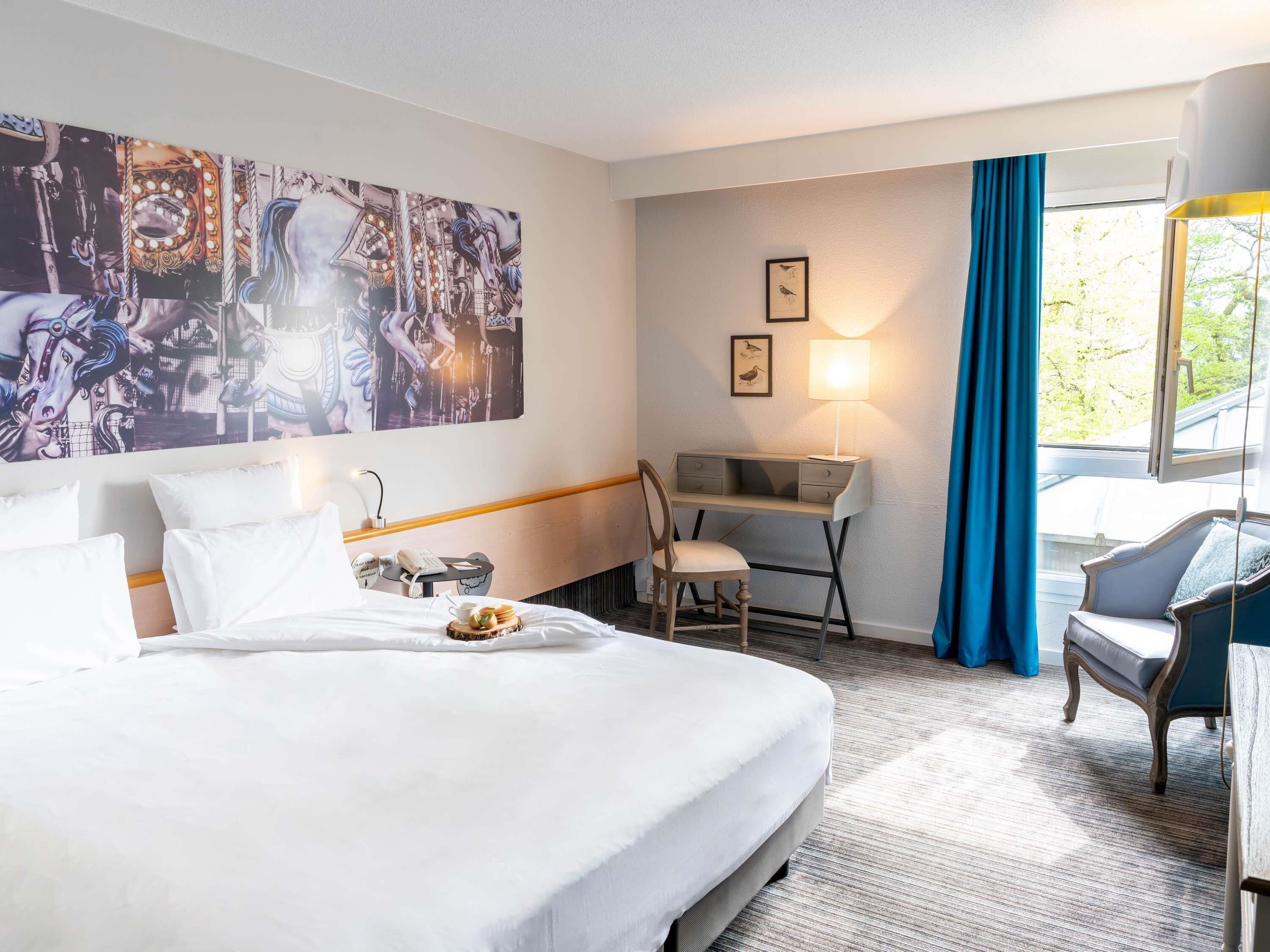 A hotel room with a large bed, a desk, a chair, a lamp, and a window with curtains.