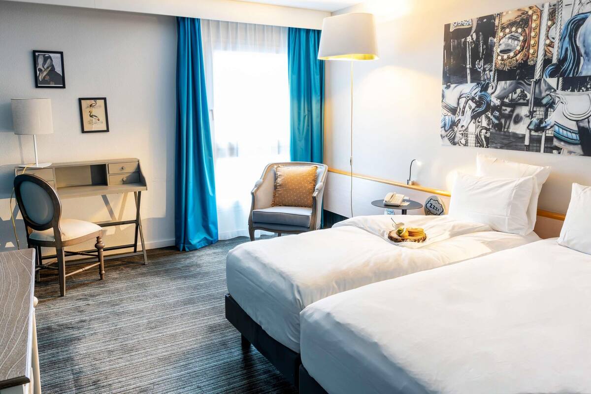 A hotel room with a bed, a desk, a chair, a lamp, and a painting on the wall.