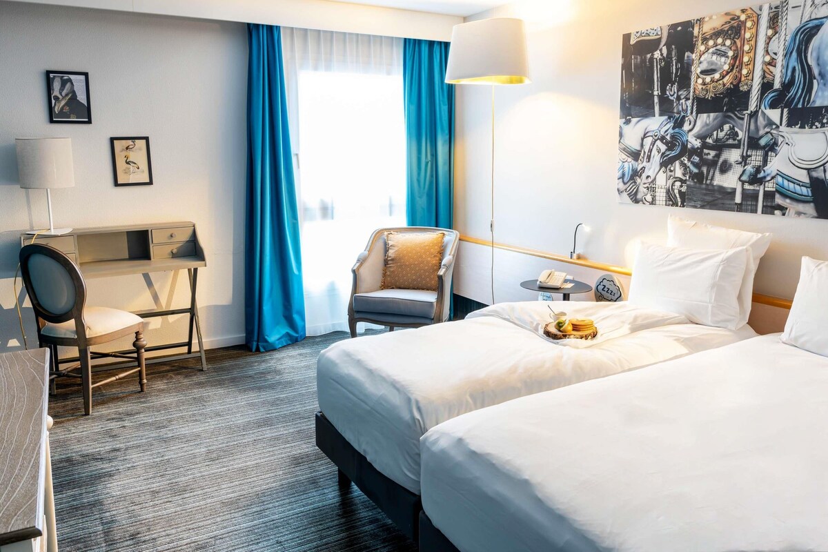 A hotel room with a bed, a desk, a chair, a lamp, and a painting on the wall.
