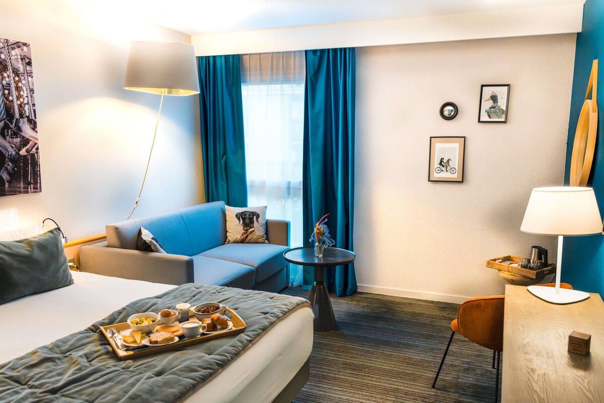 A hotel room with a bed, a sofa, a small table, a chair, a lamp, and a tray of breakfast items.