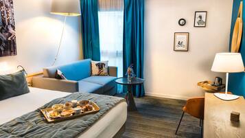 A hotel room with a bed, a sofa, a small table, a chair, a lamp, and a tray of breakfast items.