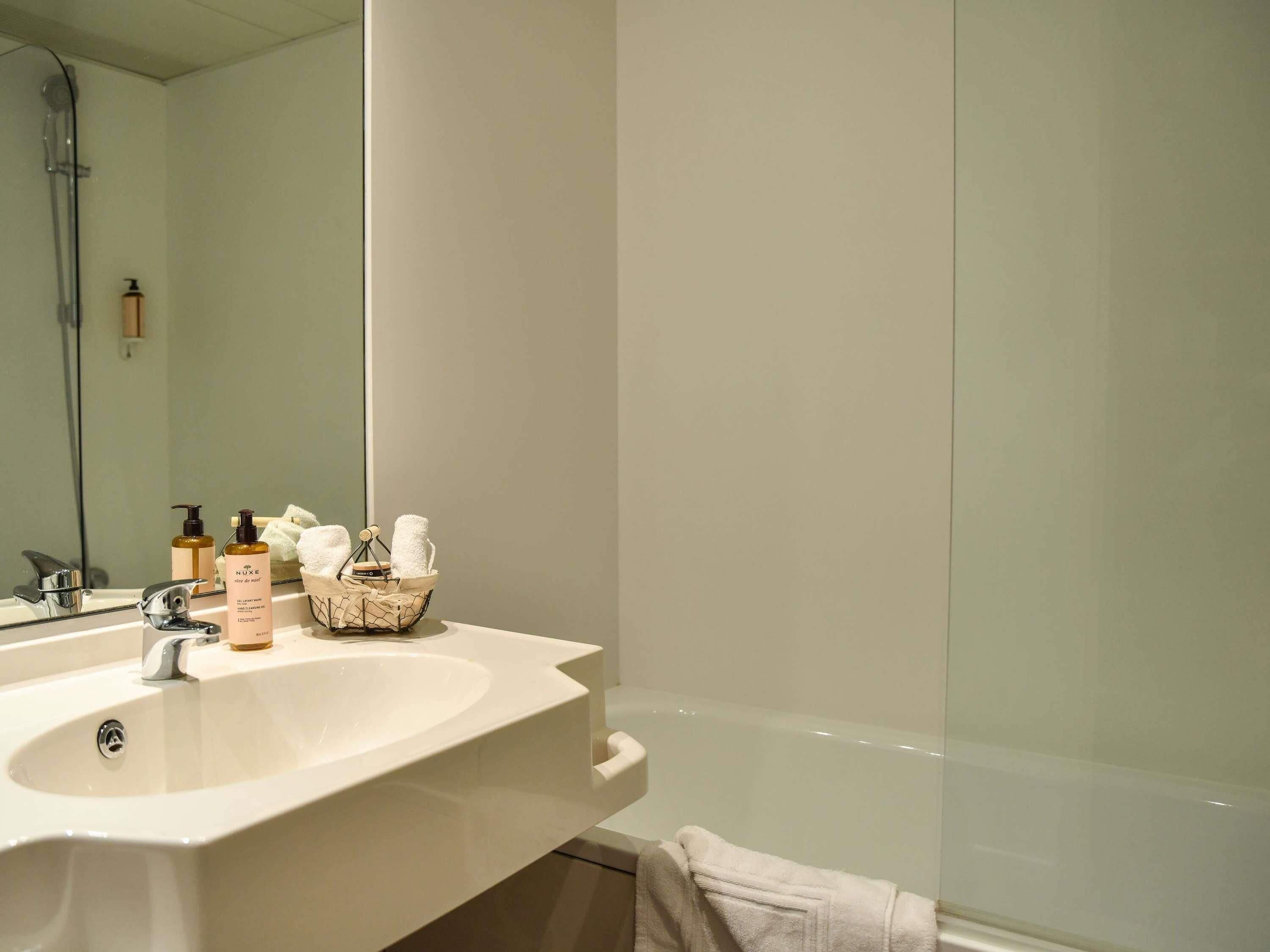 A bathroom with a white sink, a mirror, and a bathtub. There are toiletries on the sink and a towel rack with towels.