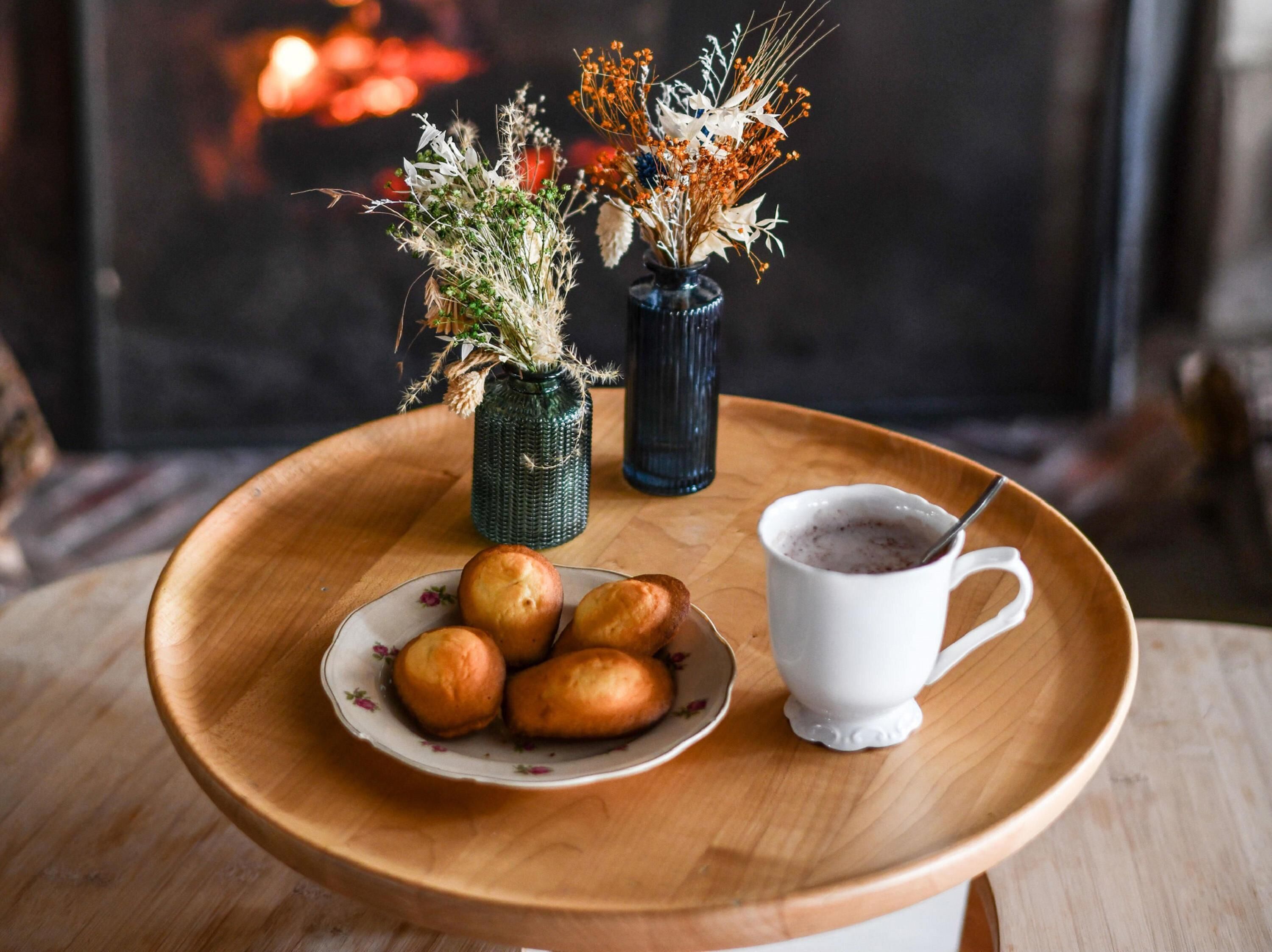 A wooden tray with a plate of round pastries, a white mug with a spoon, and two vases with dried flowers.