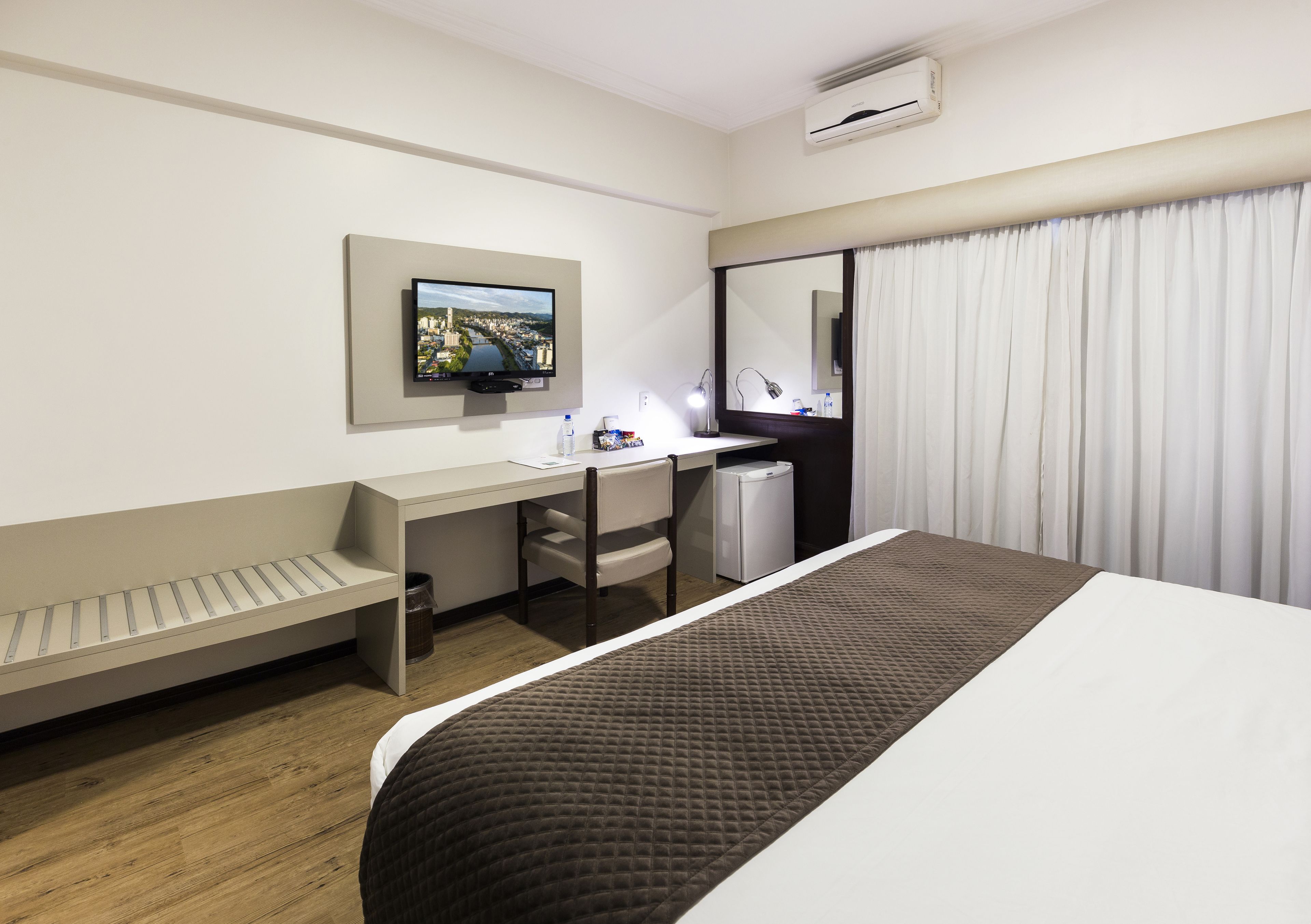 Habitación de hotel con cama, escritorio con silla, una mesita, televisor montado en la pared, ventana con cortinas y un cubo de basura.