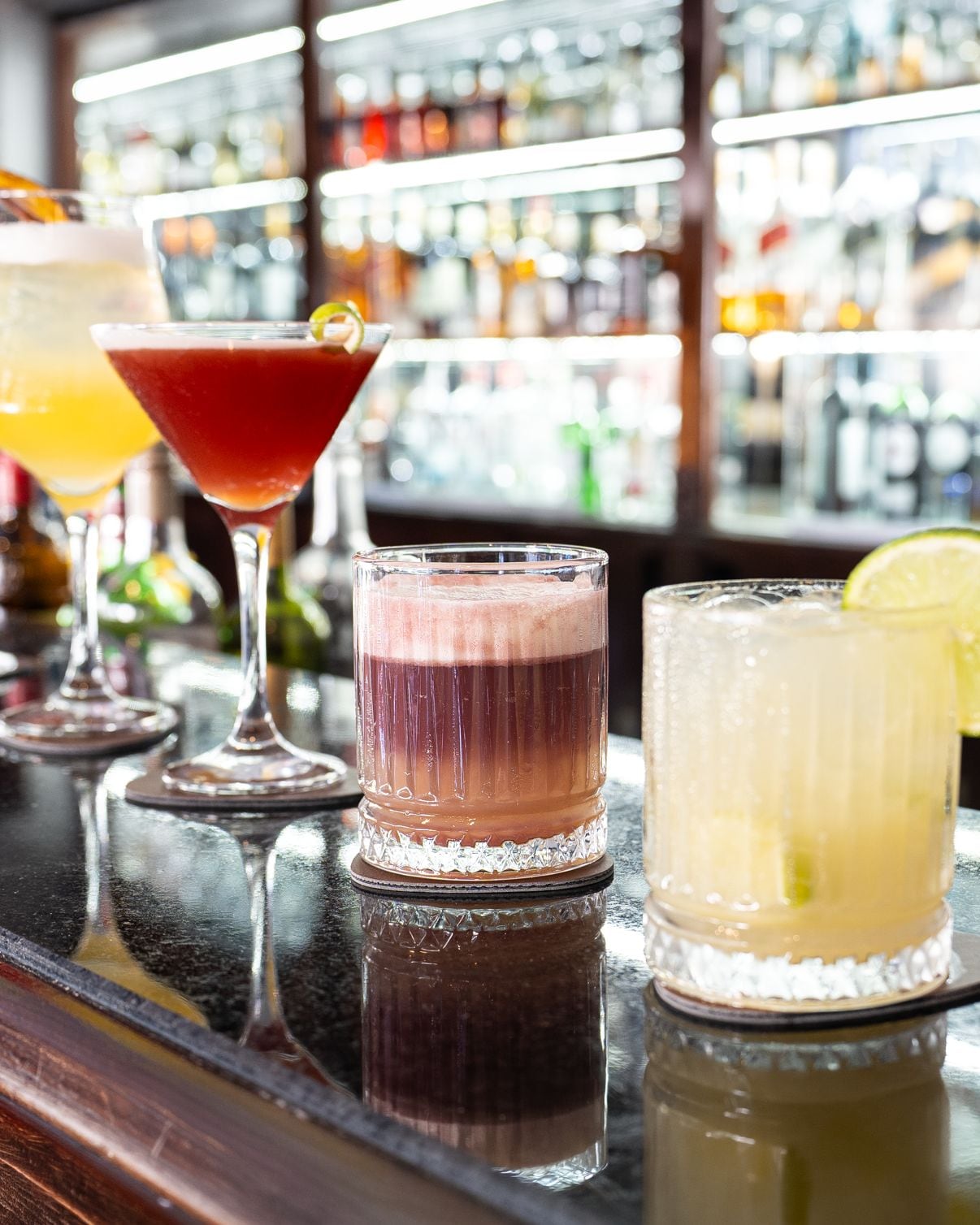 Un bar con diversas bebidas en vasos, una selección de botellas al fondo y una superficie reflectante debajo.