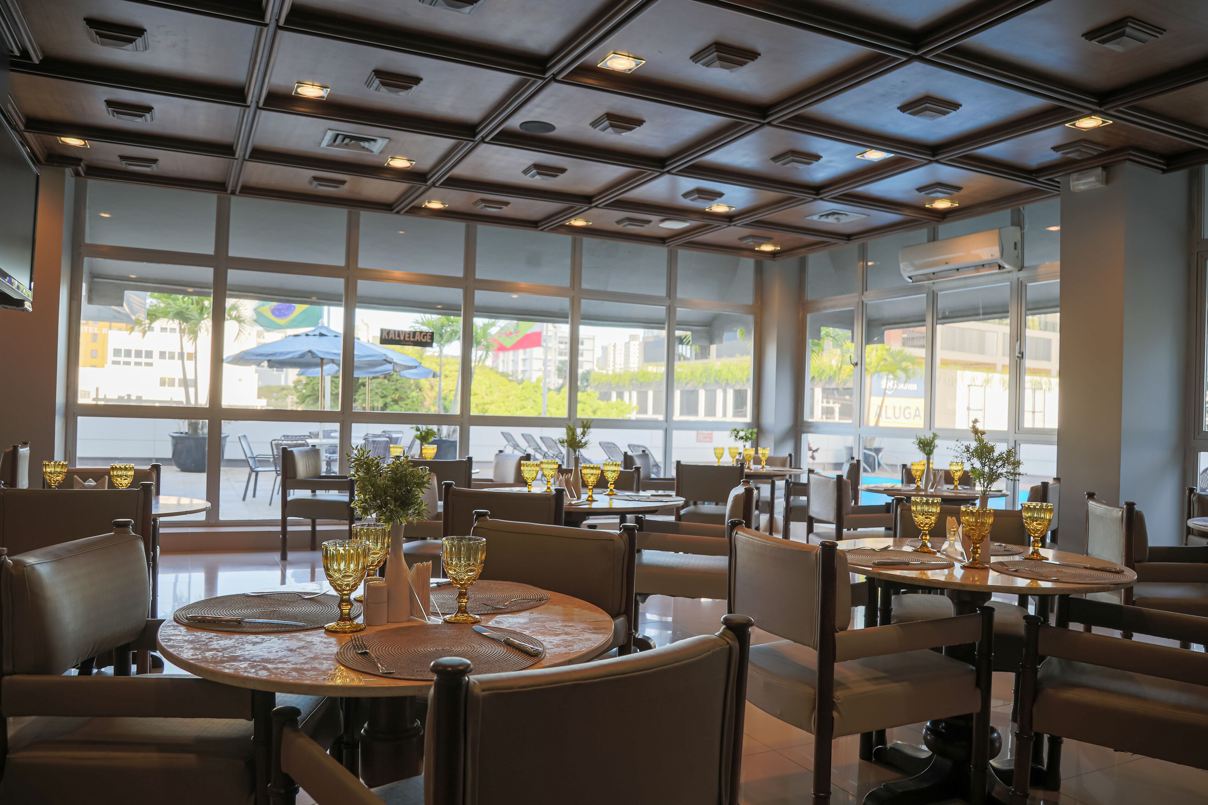 A restaurant with large windows, tables set with golden goblets, and a modern ceiling design.
