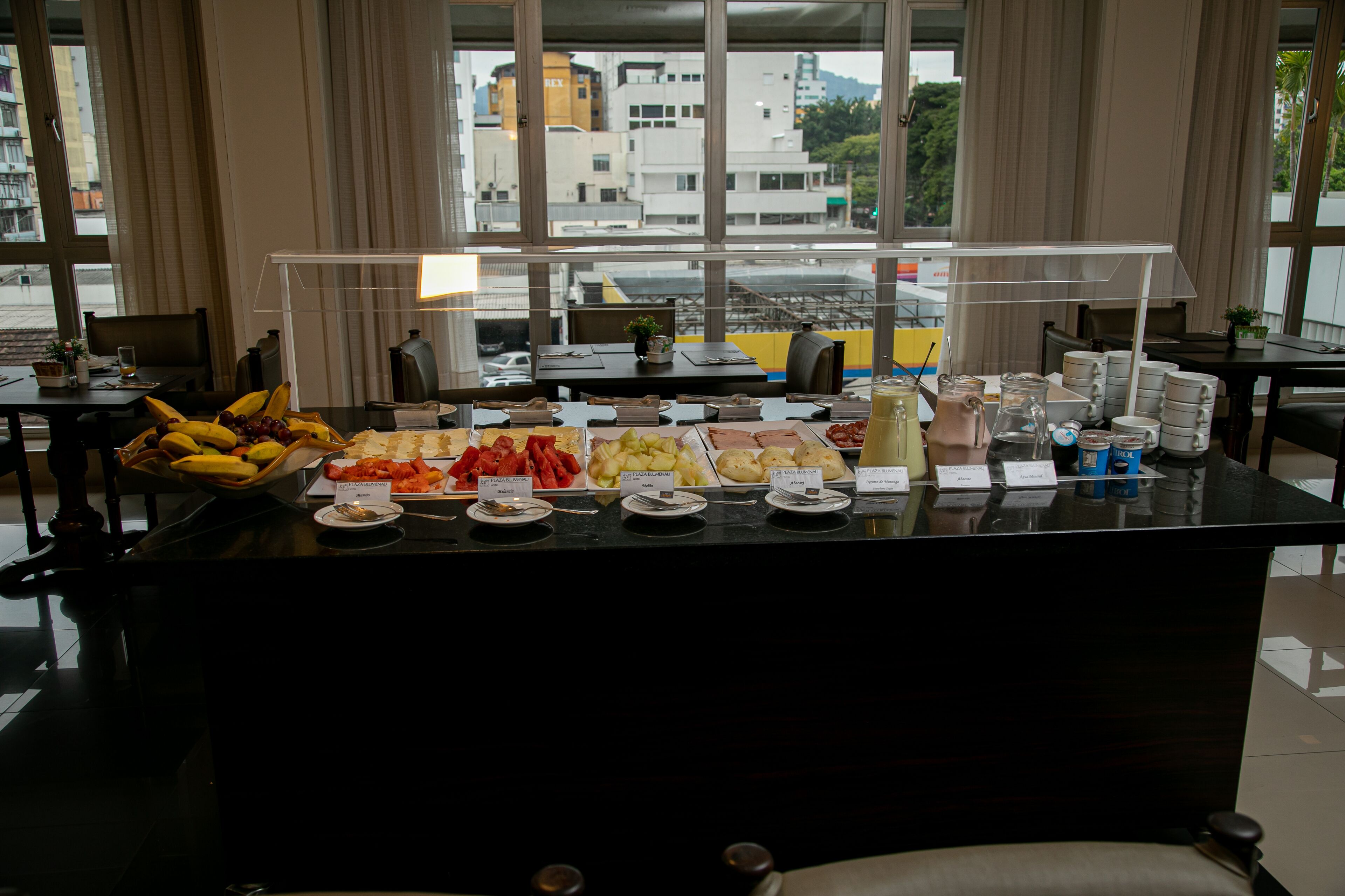 Un buffet con frutta e piatti vari, un angolo caffè e vista sulla città attraverso ampie finestre.