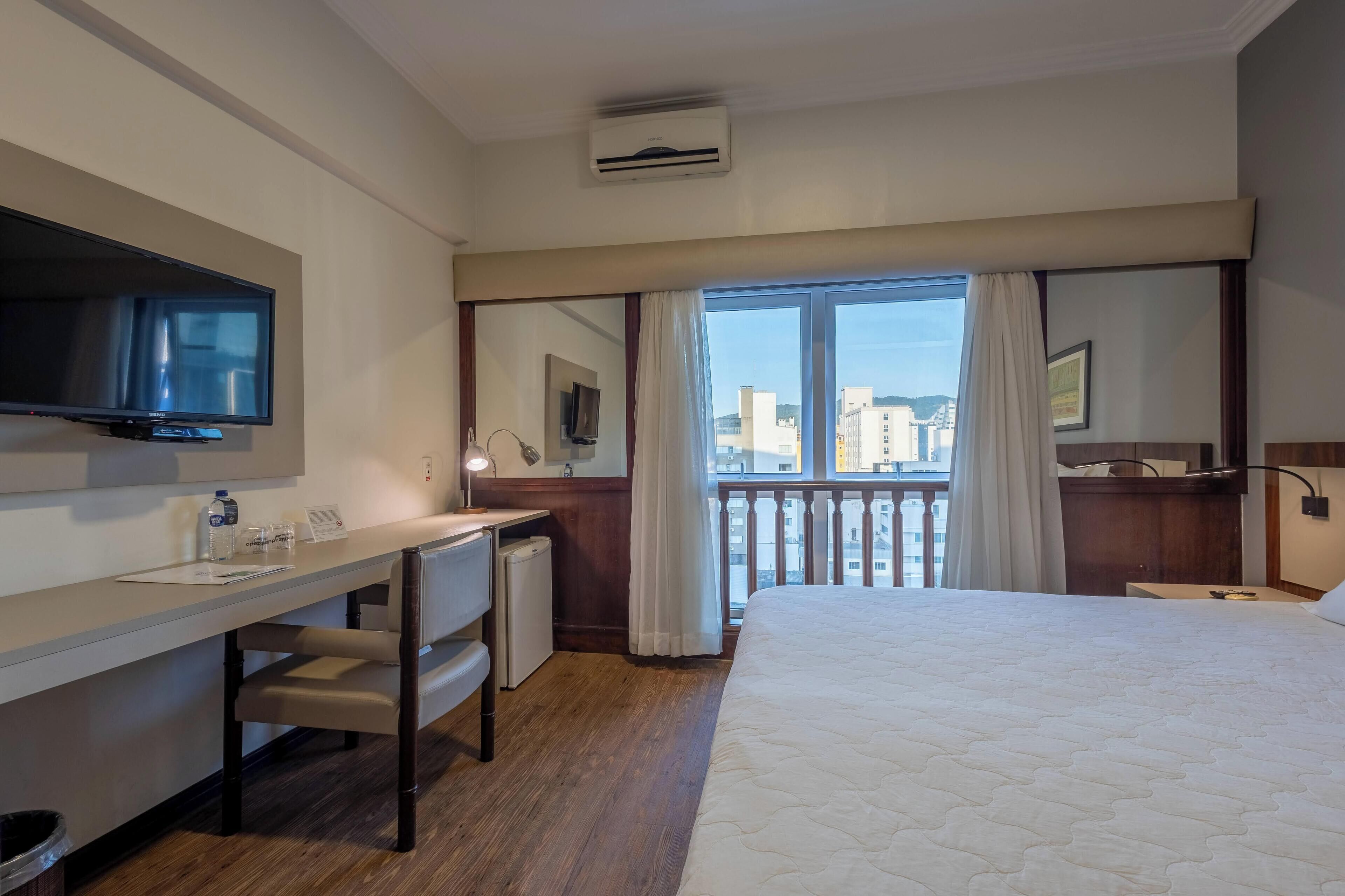 Una camera d'albergo con un letto, una scrivania, una sedia, TV, condizionatore d'aria e un balcone con tende.
