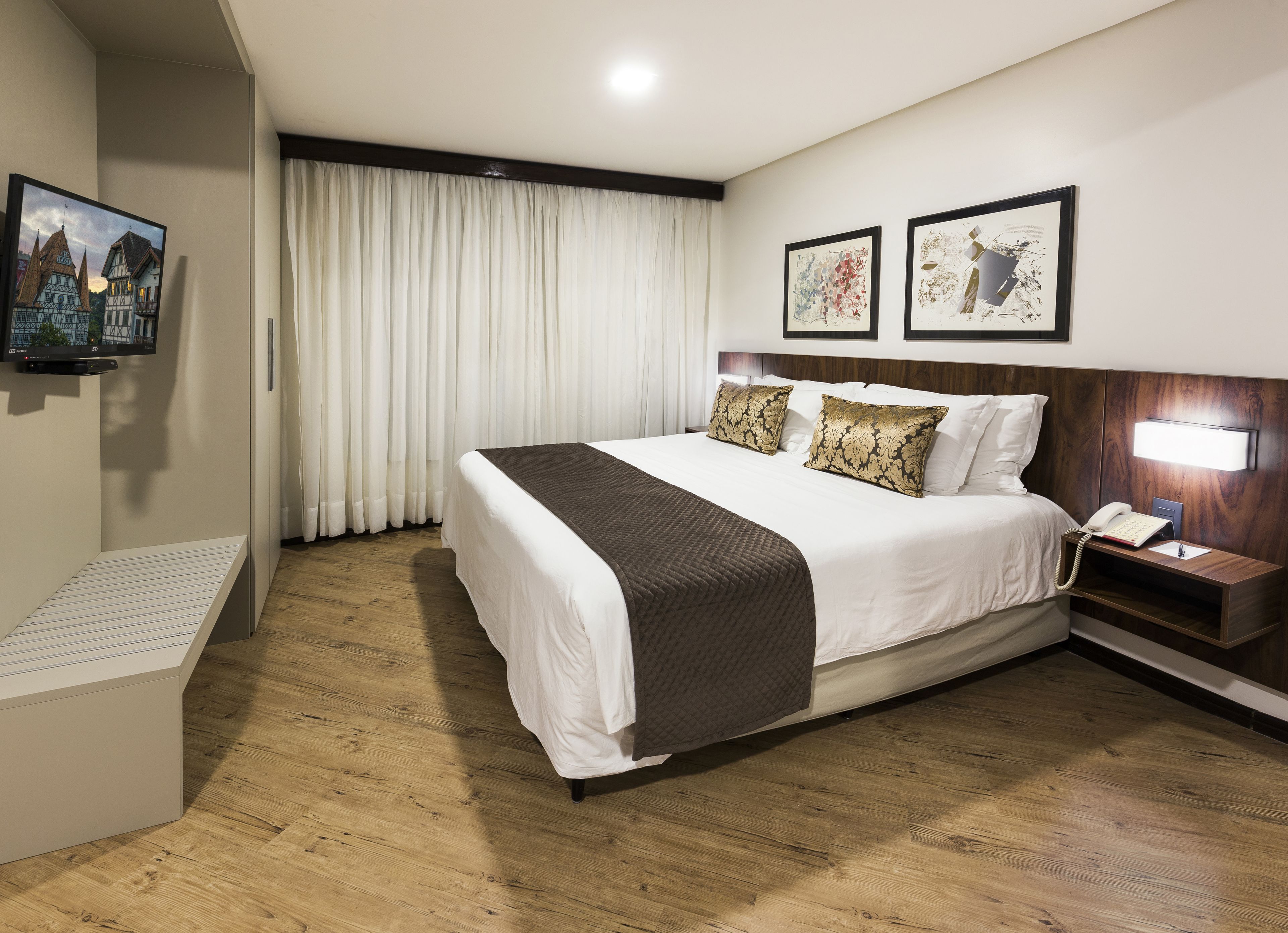 Quarto de hotel com uma cama grande, mesa de cabeceira com abajur, televisão fixada na parede e quadro emoldurado acima da cama.