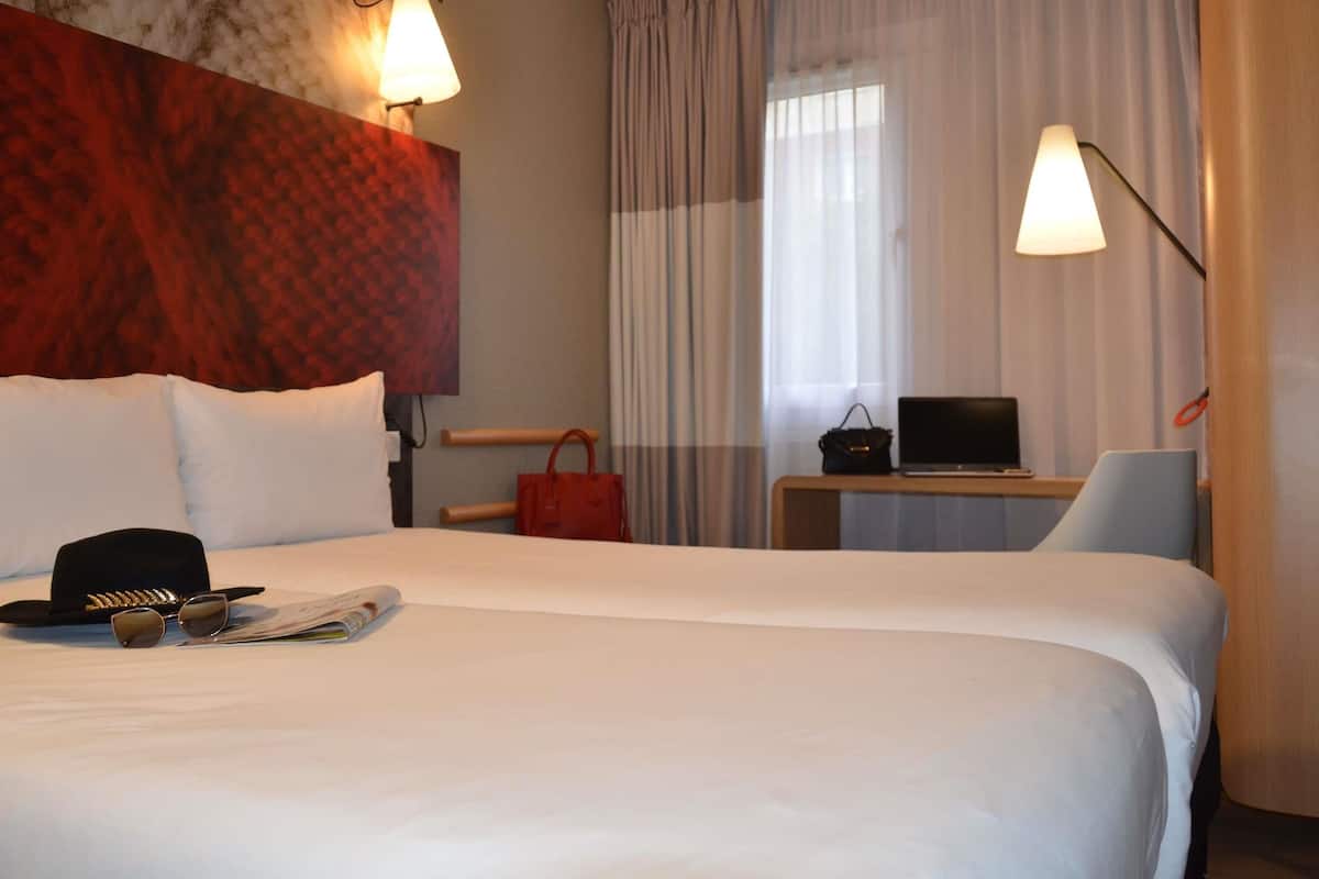 A hotel room with a large bed, a desk with a laptop, a chair, a red handbag, and a wall with a textured pattern.