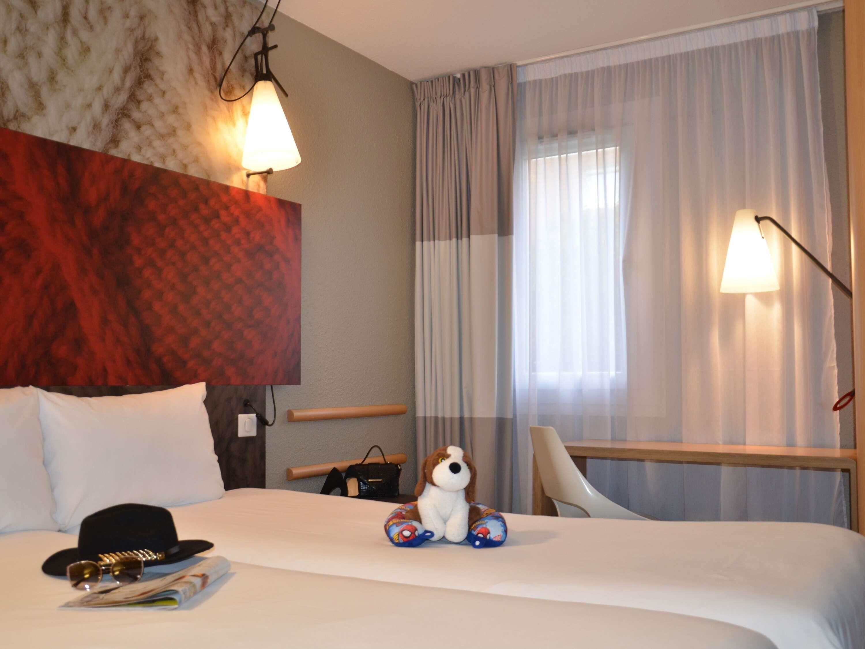 A hotel room with a bed, a headboard, a nightstand, a lamp, a window with curtains, and a plush toy.