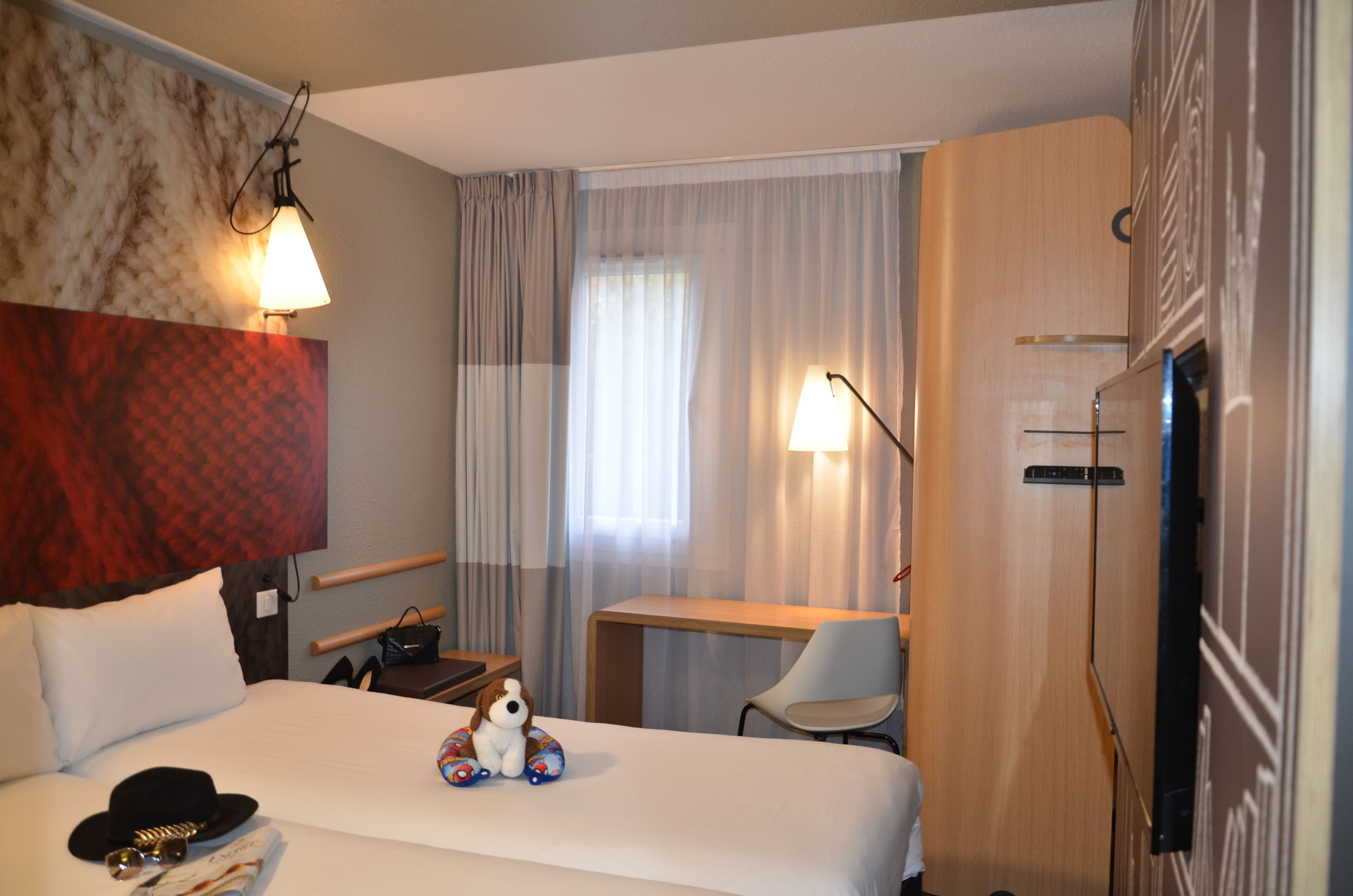 A hotel room with a bed, a desk, a chair, a TV, and a wall-mounted lamp.