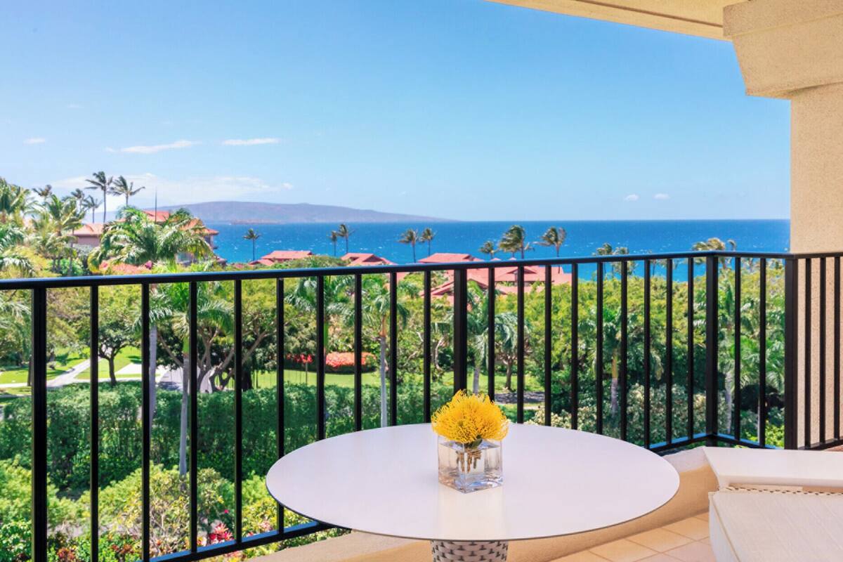 Un balcon avec une table ronde et un vase rempli de fleurs jaunes, d’où l’on peut admirer un paysage côtier avec des maisons et des palmiers.